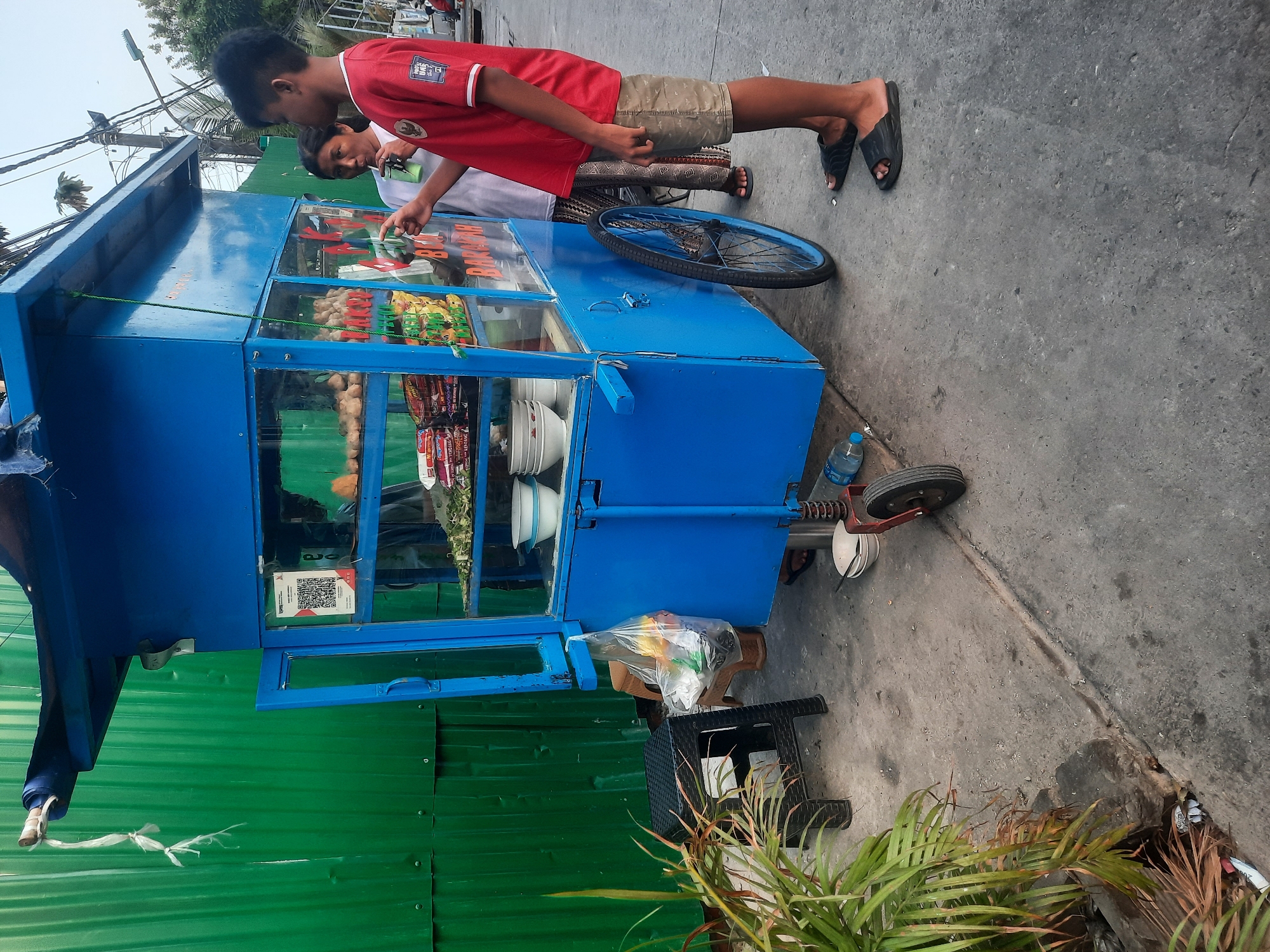 The image shows a blue food cart with a clear glass front, parked on a paved sidewalk next to a green corrugated metal fence. The cart is stocked with various food items, including bags of snacks and what appear to be meatballs on display. Two individuals, a boy wearing a red shirt and shorts, and a woman wearing a white t-shirt and patterned pants, are standing near the cart. The woman is holding a green cup with a straw and drinking from it. The boy is pointing towards the cart, possibly interacting with the woman or the cart's contents. The environment suggests an urban setting, likely in North Jakarta, Indonesia, given the presence of tropical plants in the foreground and electrical wires overhead. The overall scene depicts a typical street-side food vendor operation.