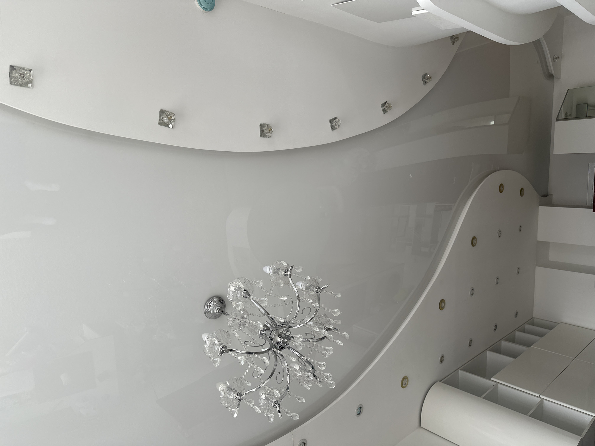 The image depicts the interior of a room, possibly located in Khimki, Russia, featuring a decorative ceiling with a modern design. The ceiling is predominantly white, with curved, layered sections and embedded square crystal-like accents. A chandelier with multiple glass or crystal elements is centrally positioned. The room appears modern and well-lit, focusing on ceiling details and built-in storage. The room is likely a bedroom or a living area with a custom-designed interior.