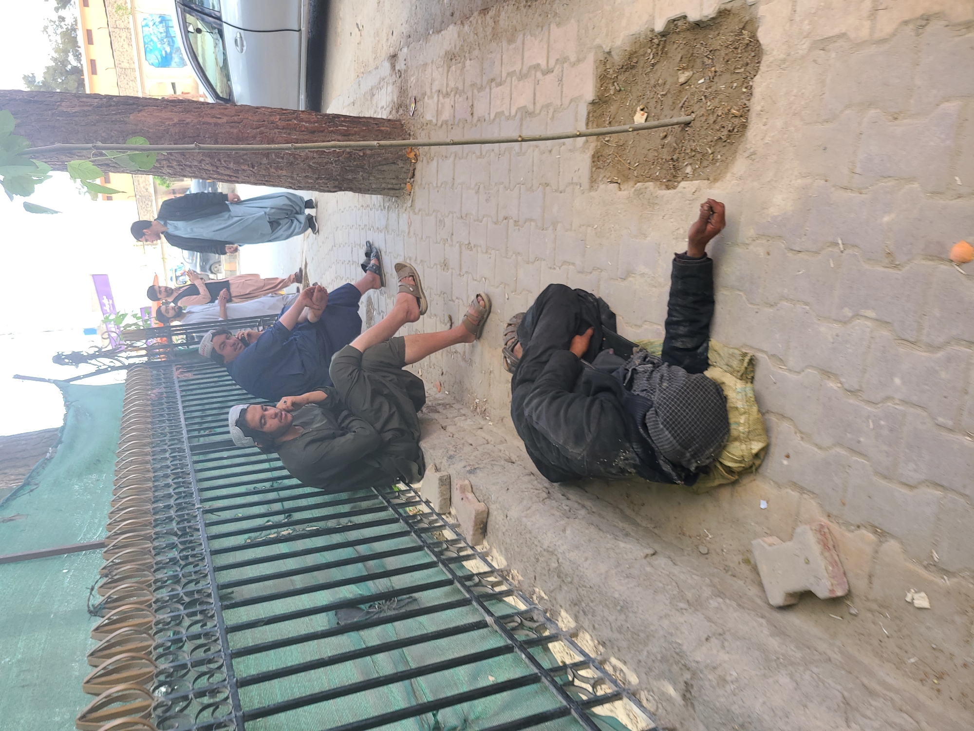 The image shows a group of men lying on a sidewalk next to a fence and a tree, likely in Quetta City Tehsil, Pakistan. One man is lying on his side on a tarp, dressed in dark clothing. Three other men are reclining against the fence, one in a blue outfit, one in green, and another further back in a beige garment. A standing man in light blue is visible behind them, next to a parked car. The sidewalk is paved with interlocking bricks, and there's an unpaved patch with dirt next to it.