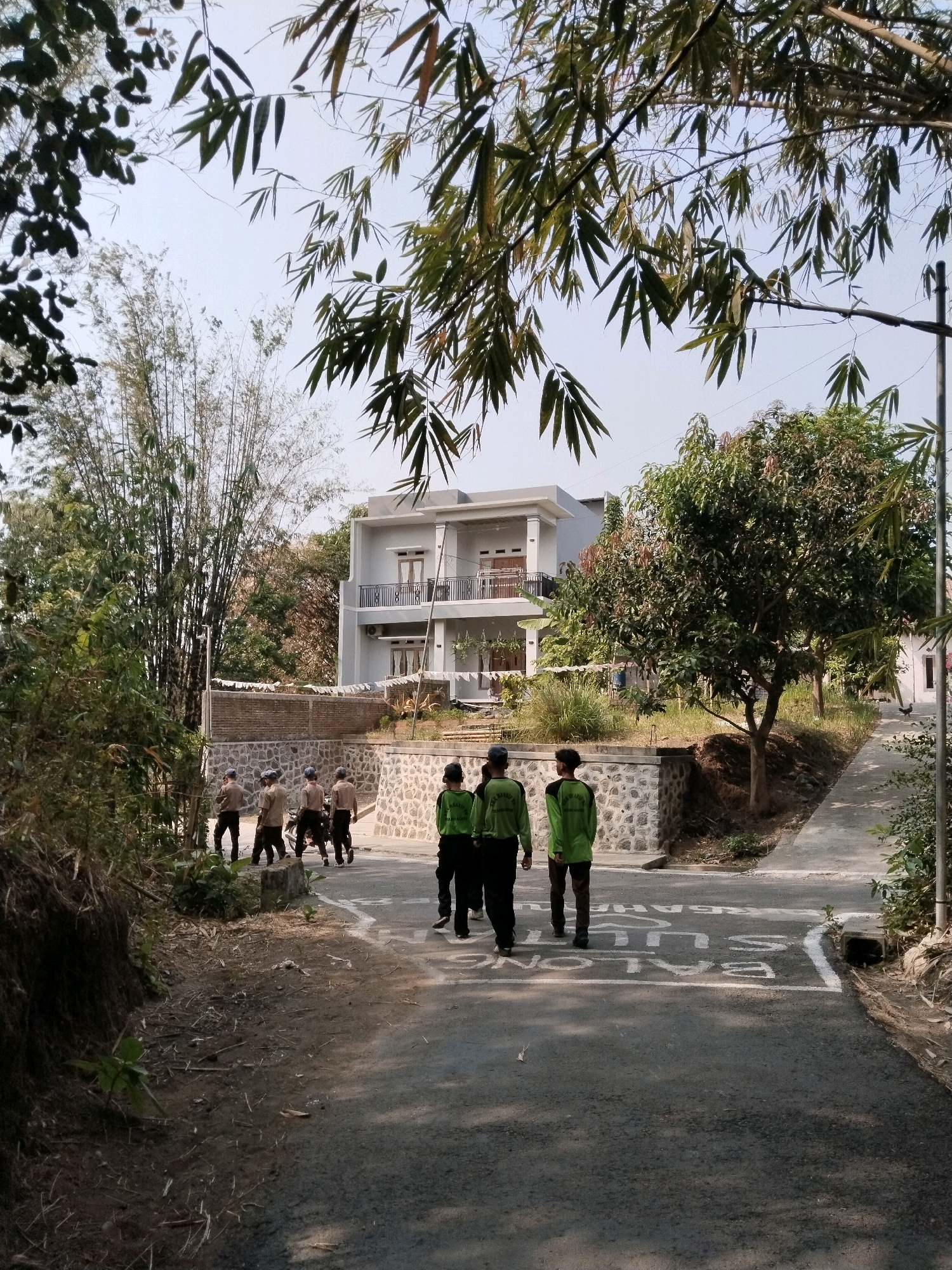 The image depicts a street scene in Pakulaut, Indonesia, featuring a group of individuals in uniforms walking away from the camera on an asphalt road. The road has white markings that appear to be part of a traffic or safety instruction, with text "SELALU BALONGSULIT" visible.
In the foreground, a slope covered in dry leaves and sparse vegetation is on the left side of the road. The road itself is shaded in some areas, suggesting it's daytime with the sun casting shadows.
Behind the walking group, there is a stone wall topped with a low brick wall, bordering a raised area. On this raised area, several trees are planted, including a prominent leafy tree with reddish-tinged new growth. Beyond the trees, a modern, two-story house with a light-colored facade and balconies stands on a higher elevation.
To the left of the house, a dense stand of tall bamboo trees is visible, adding to the lush, natural surroundings. The sky is clear and bright, indicating fair weather.
The individuals are dressed in uniforms. One group of four appears to be wearing light brown shirts and dark pants, possibly representing a scout or school uniform. Another group of two, closer to the foreground, are wearing bright green long-sleeved shirts with dark pants.
The overall impression is a serene, possibly educational or community setting in a semi-rural or suburban part of Pakulaut, Indonesia.
