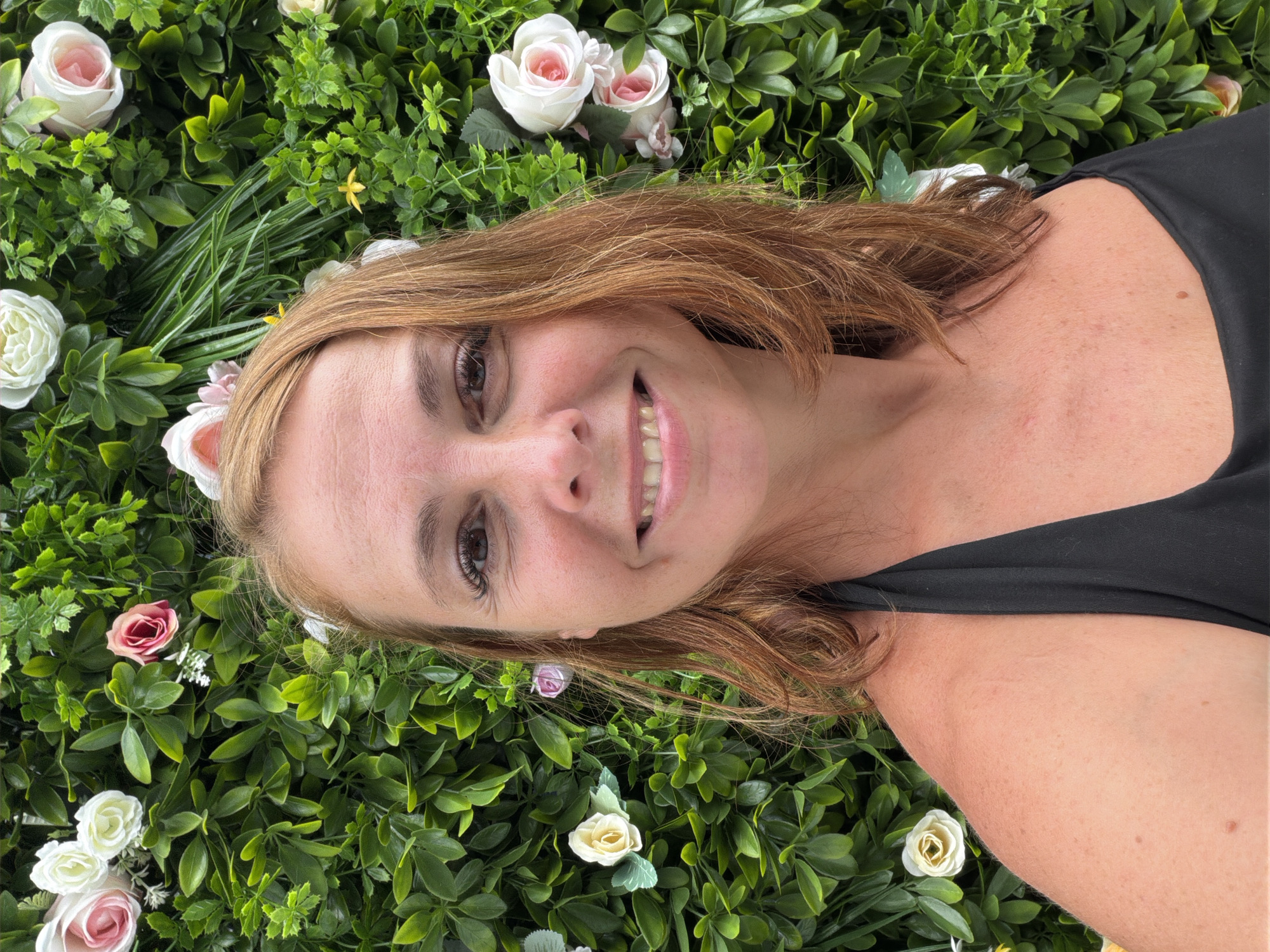 In the city of Ban Rang Rak, Thailand, a woman with auburn hair and a black halter top smiles at the camera, positioned in front of a lush green floral backdrop. The backdrop features an abundance of artificial greenery interspersed with white, pink, and yellow roses of various sizes. Her face is illuminated by frontal lighting, accentuating her smile and the details of her makeup. She is wearing earrings and her hair is styled to fall around her shoulders. The image captures a close-up portrait, focusing on the woman and the vibrant floral arrangement behind her.