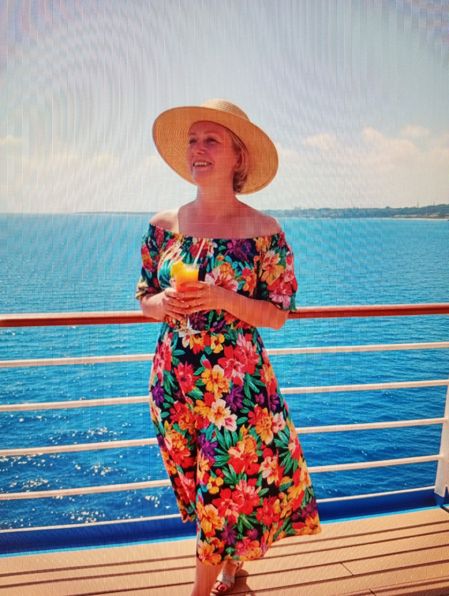 A full shot captures a Caucasian woman standing on a ship deck with a bright orange juice in her hand. She is wearing a straw hat and an off-the-shoulder dress with a floral pattern. The woman is looking to her right, with a smiling expression. The background shows a bright blue sea and a hazy sky.
