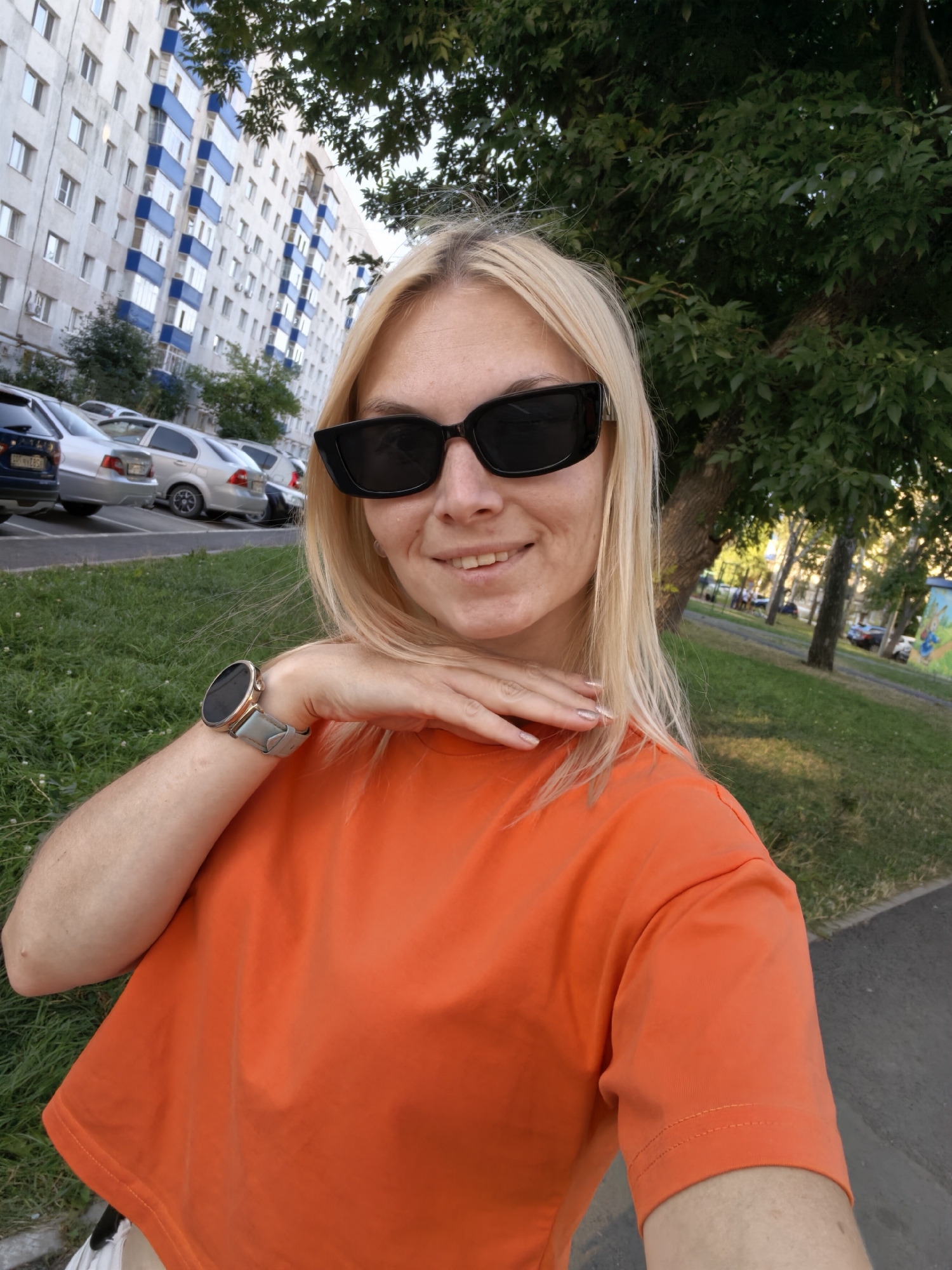 A blonde woman with sunglasses and an orange t-shirt is standing outside a building, possibly an apartment complex, with cars parked in the background. She is holding her hand to her neck. The location is the city of Sterlitamak, Russia. The image appears to be a selfie taken in daylight.