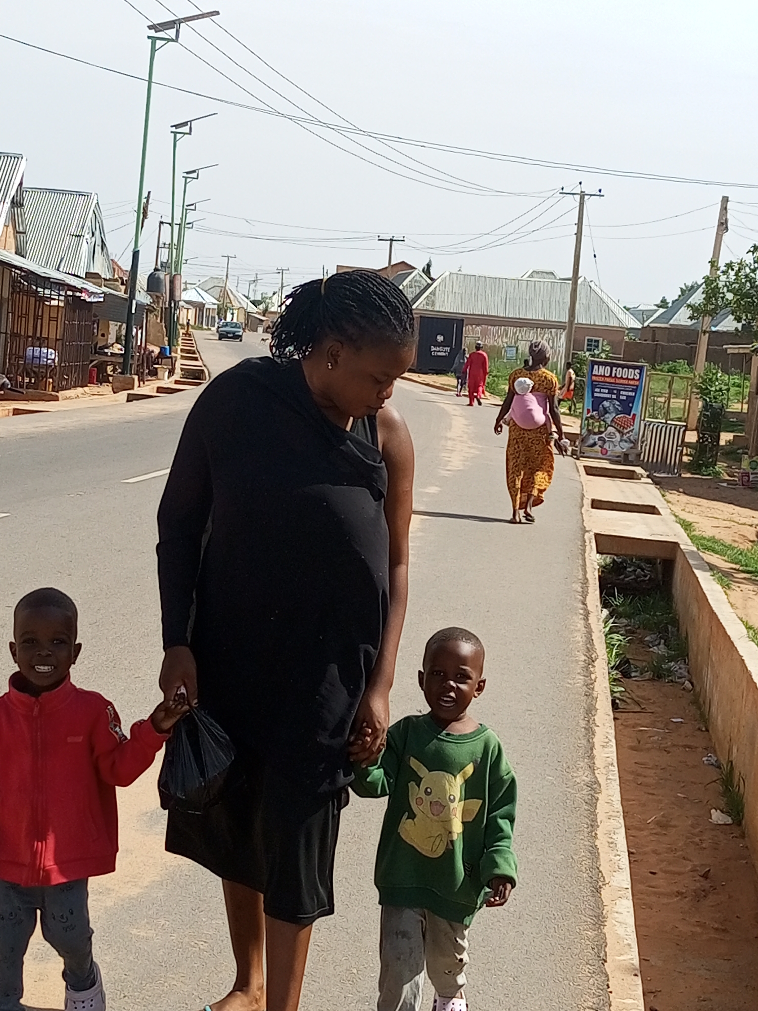 The image depicts a street scene in Tempure, Nigeria. In the foreground, a woman is walking with two young children. She is wearing a black outfit and carrying a black plastic bag. The child on her left is wearing a red jacket and jeans, and the child on her right is wearing a green sweatshirt with a Pikachu design and grey pants. They are all walking on an asphalt road. In the background, there are buildings with corrugated iron roofs, solar-powered streetlights, and power lines. A woman carrying a baby on her back is walking further down the road, and another woman in a yellow dress is walking in the same direction. There is also a sign for "ANO FOODS." The environment appears to be a typical urban or semi-urban setting with some vegetation along the roadside.