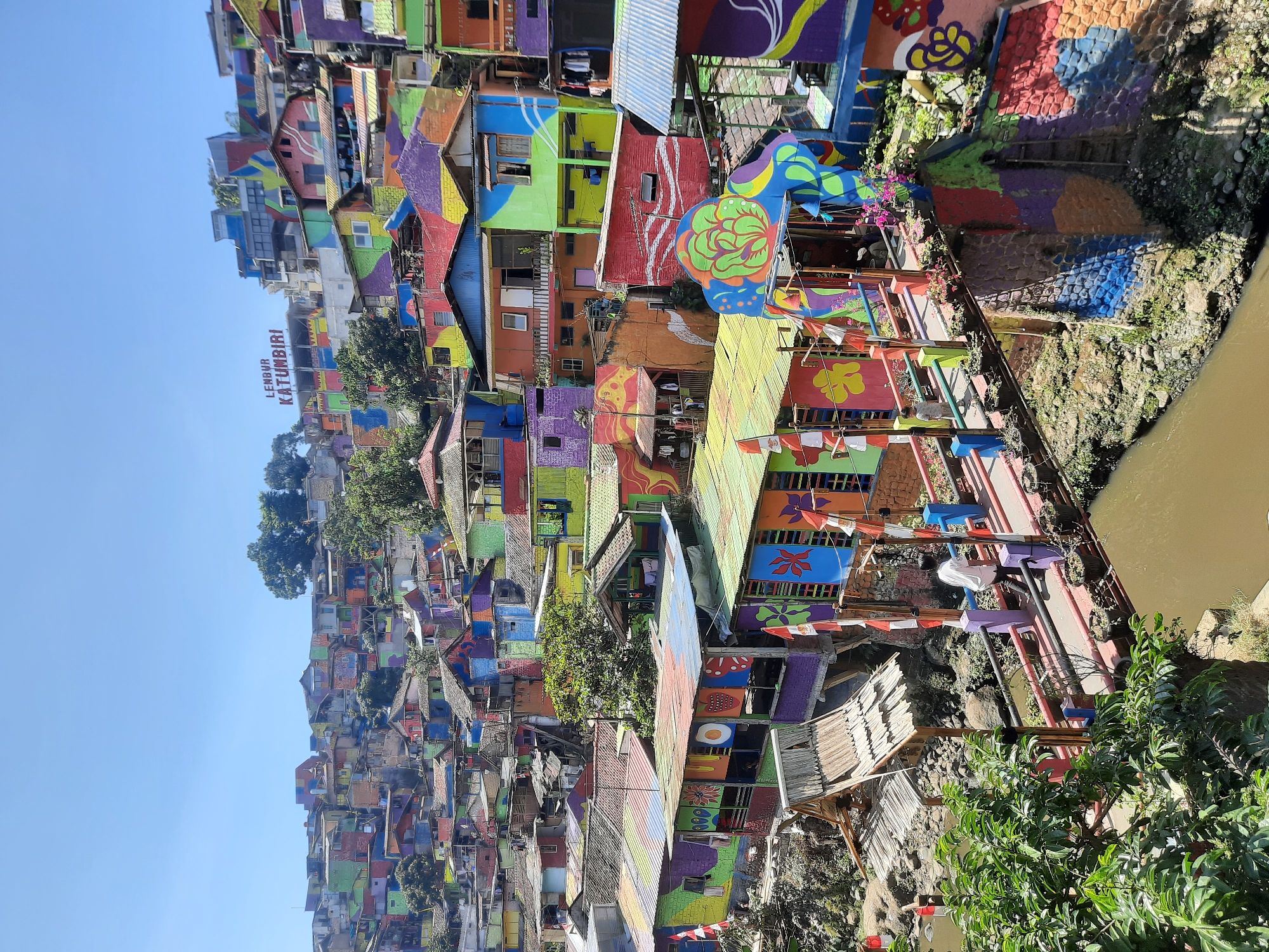 The image depicts a vibrant, densely populated hillside settlement in Bandung City, Indonesia, known as Kampung Pelangi or "Rainbow Village." The houses, densely packed and built on a steep incline, are painted in a riot of bright, contrasting colors, including red, blue, green, yellow, purple, and orange. Many of the houses feature intricate murals and decorative elements. A wooden bridge, also adorned with colorful paint and flags, spans a murky river flowing through the lower part of the scene. A single person, wearing a white shirt, walks across the bridge. Lush green foliage surrounds the lower sections of the village, contrasting with the colorful structures. The sky above is clear and blue. A sign at the top of the hill reads "LEMBUR KATUMBINI."