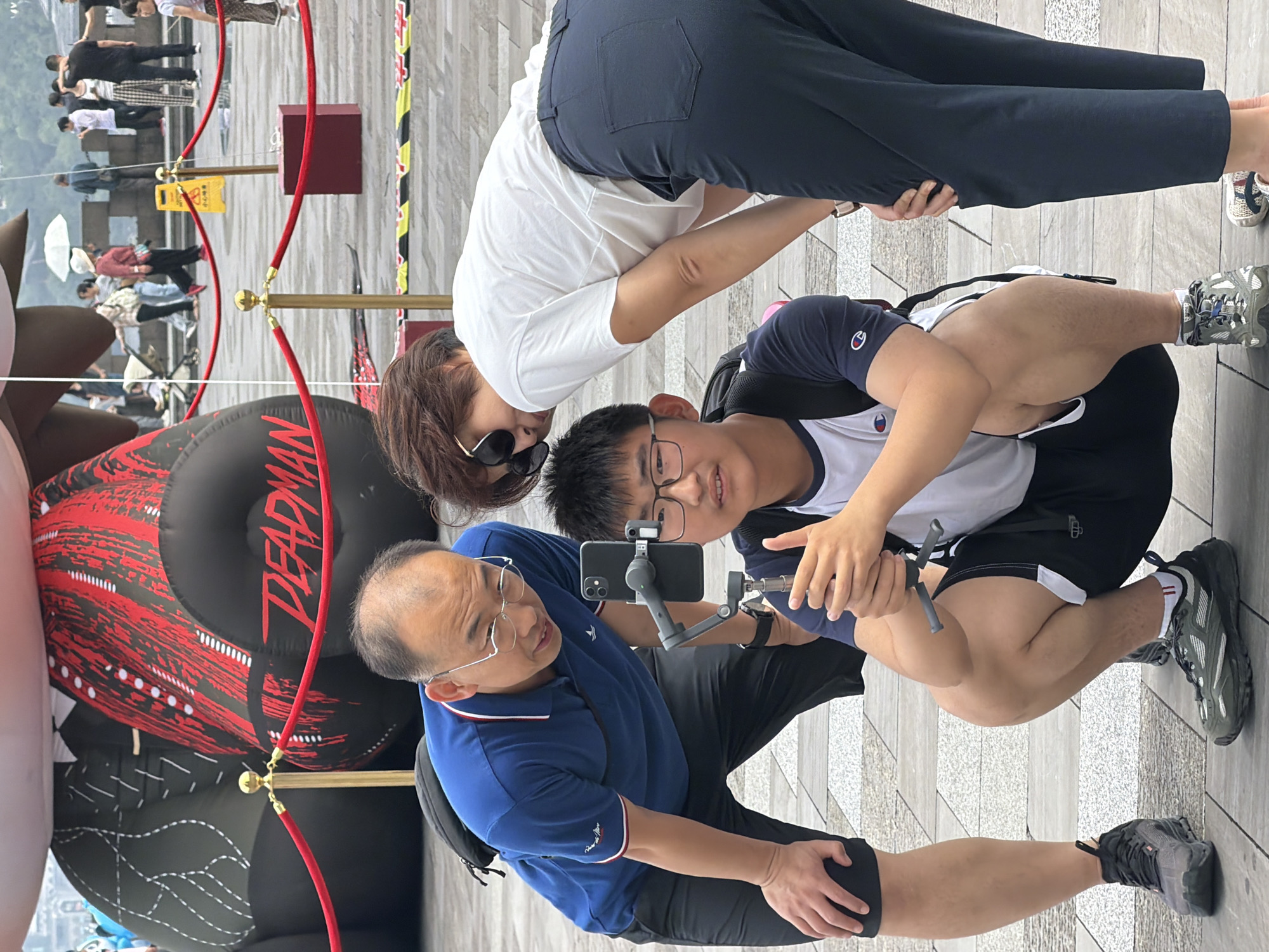 In Yuzhong District, China, three individuals are gathered around a mobile phone mounted on a gimbal. A young male, wearing glasses, a white and navy t-shirt, and black shorts, is crouching and holding the gimbal with the phone attached, seemingly filming or taking pictures. He is positioned in the foreground. To his left, an adult male in a blue polo shirt and black shorts, also wearing glasses, leans forward and looks at the phone. He has a backpack slung over his shoulder. To the right of the young male, an adult female, wearing a white t-shirt and dark pants, is bent over, also looking at the phone. She has sunglasses on her head. In the background, a large inflatable structure with "DEADMAN" written on it in red is visible behind a red velvet rope barrier supported by gold stanchions. Other people and urban scenery are visible further in the background, including buildings and pedestrians. The ground is paved with square tiles.