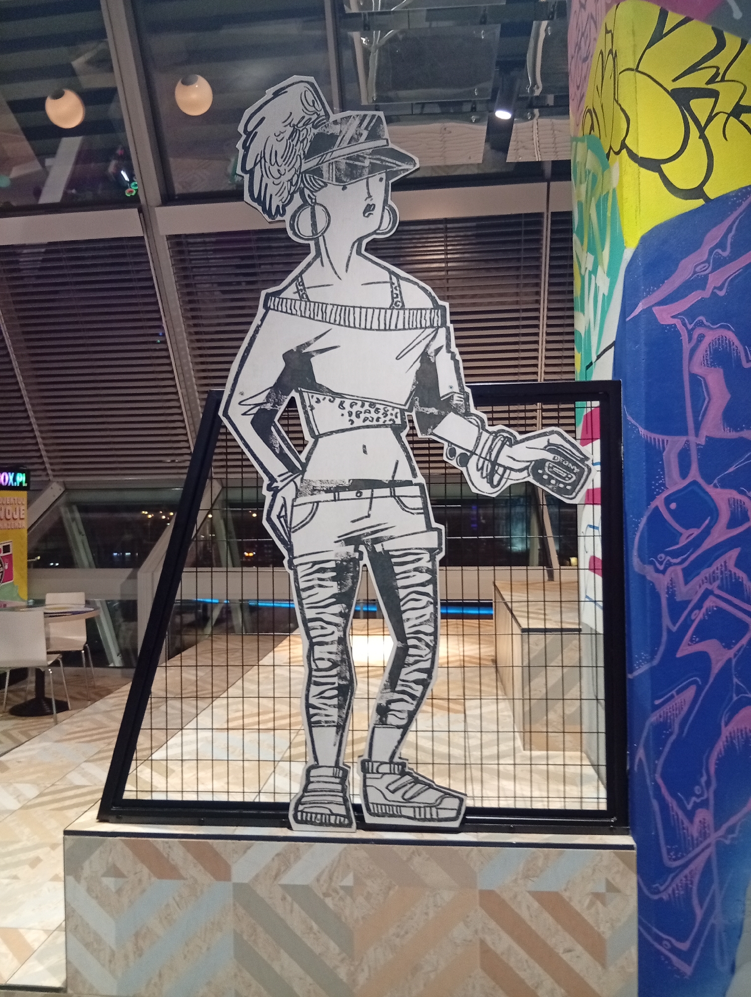 The image shows a life-size, black and white cutout of a woman wearing a hat, sunglasses, a loose-fitting top, shorts, and striped leggings, standing behind a black metal mesh fence. She is holding a portable music player in her right hand. To the right of the cutout is a vibrantly painted pillar with graffiti art in yellow, blue, and pink. In the background, there is a large window with a view of lights and the interior of a building with tables and chairs. The floor is tiled with a geometric pattern. The scene is located at the Sky Tower in Wrocław, Poland.