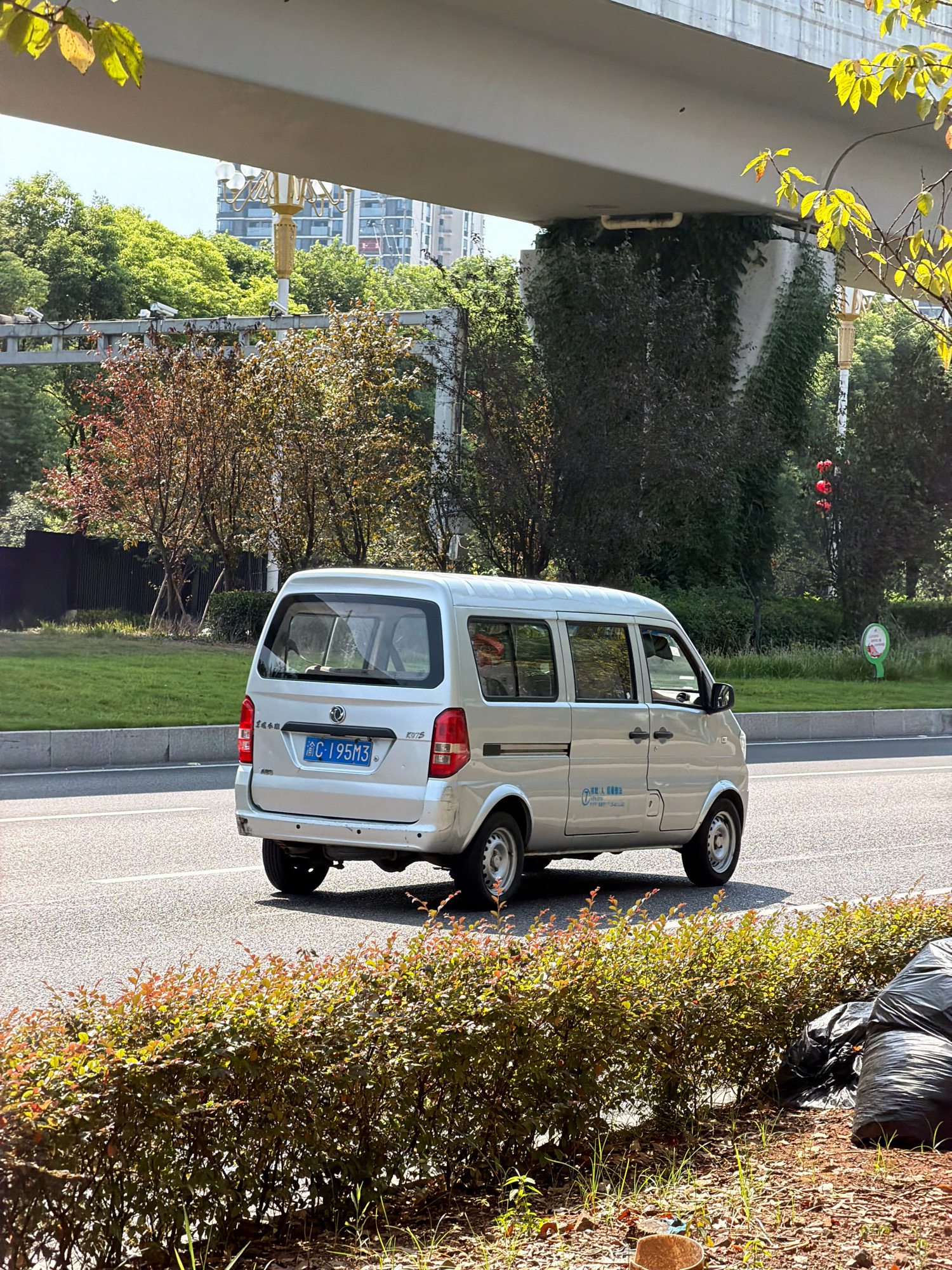 A silver minivan is driving on a road in Jiangjin, China. The minivan has a license plate that reads "C·195M3". The road is lined with trees and bushes. In the background, there are buildings and an overpass. The weather is sunny. There are no people visible in the image.