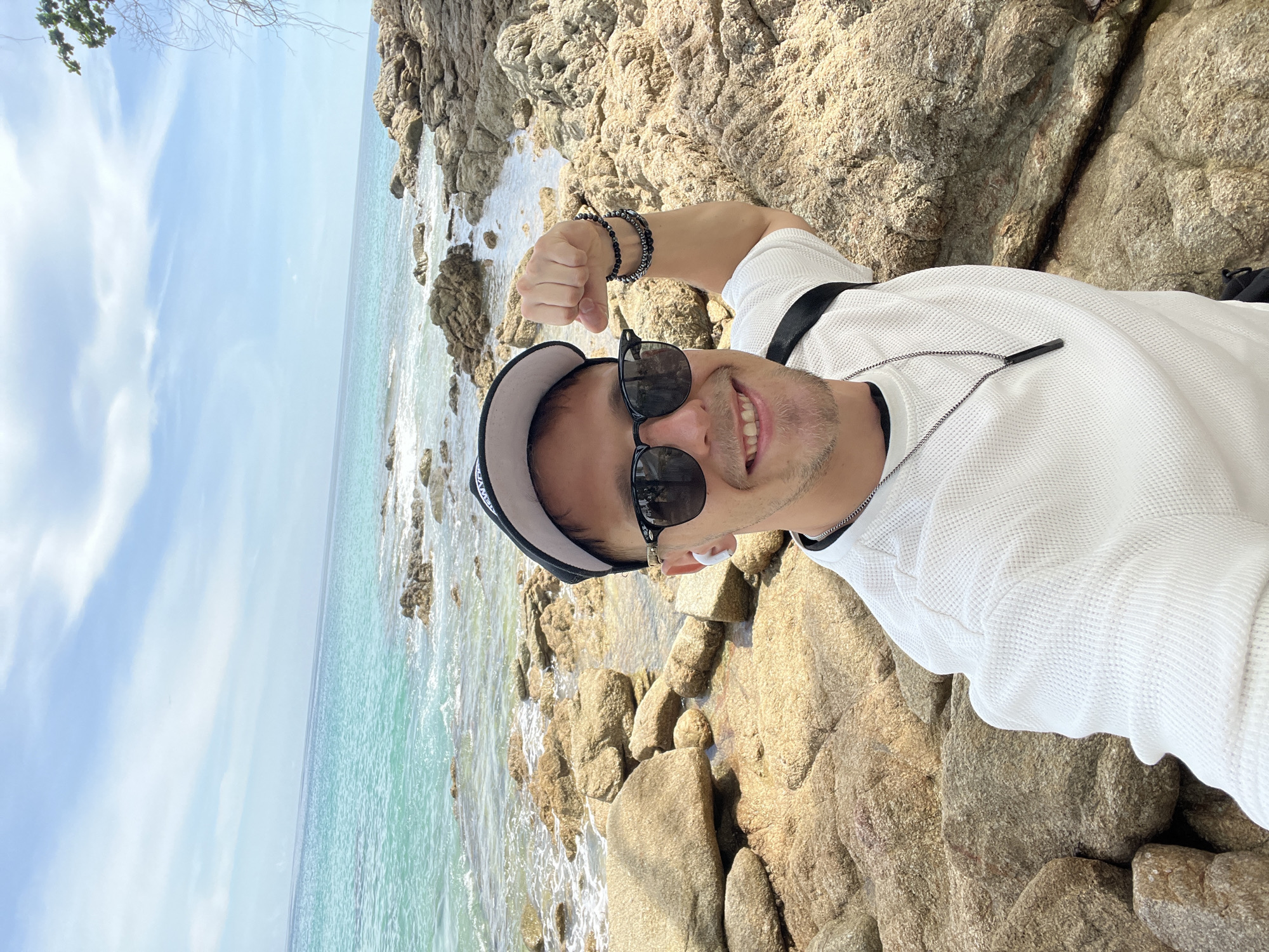 A man is taking a selfie on a rocky beach. He is wearing a white waffle-knit shirt, a black baseball cap, and sunglasses. He has two bracelets on his left wrist and a necklace with a black pendant. The man is smiling and flexing his bicep.
The scene is a tropical beach with turquoise water and large rocks. The sun is shining, indicating it is daytime. The weather appears to be warm and clear, with a few clouds in the sky. The man's attire suggests a casual setting, likely a vacation or a day trip to the beach. The image conveys a sense of happiness and enjoyment.
The location is identified as Choeng Thale, Thailand, which aligns with the tropical beach scenery. The rocks appear to be volcanic, common in some parts of Thailand. The water is clear and inviting, suggesting a warm climate. No visible text is present in the image. The overall impression is one of a relaxed and joyful individual enjoying the natural beauty of a Thai beach.