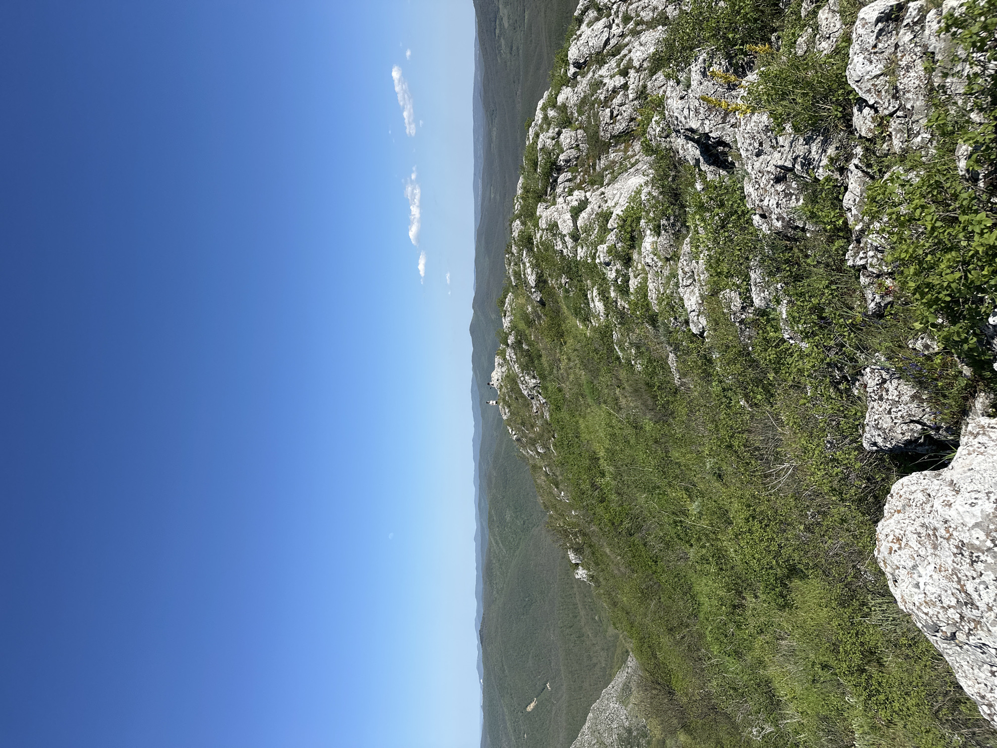 The image shows a landscape with a clear blue sky occupying the left half, contrasted by a rocky, vegetation-covered terrain on the right. The landscape features a ridge or edge of a mountain, characterized by light-colored rocks and interspersed with green plants. The view extends to a range of hills in the distance, appearing somewhat hazy. The photo appears to be taken on a sunny day in Bilka, Bulgaria.