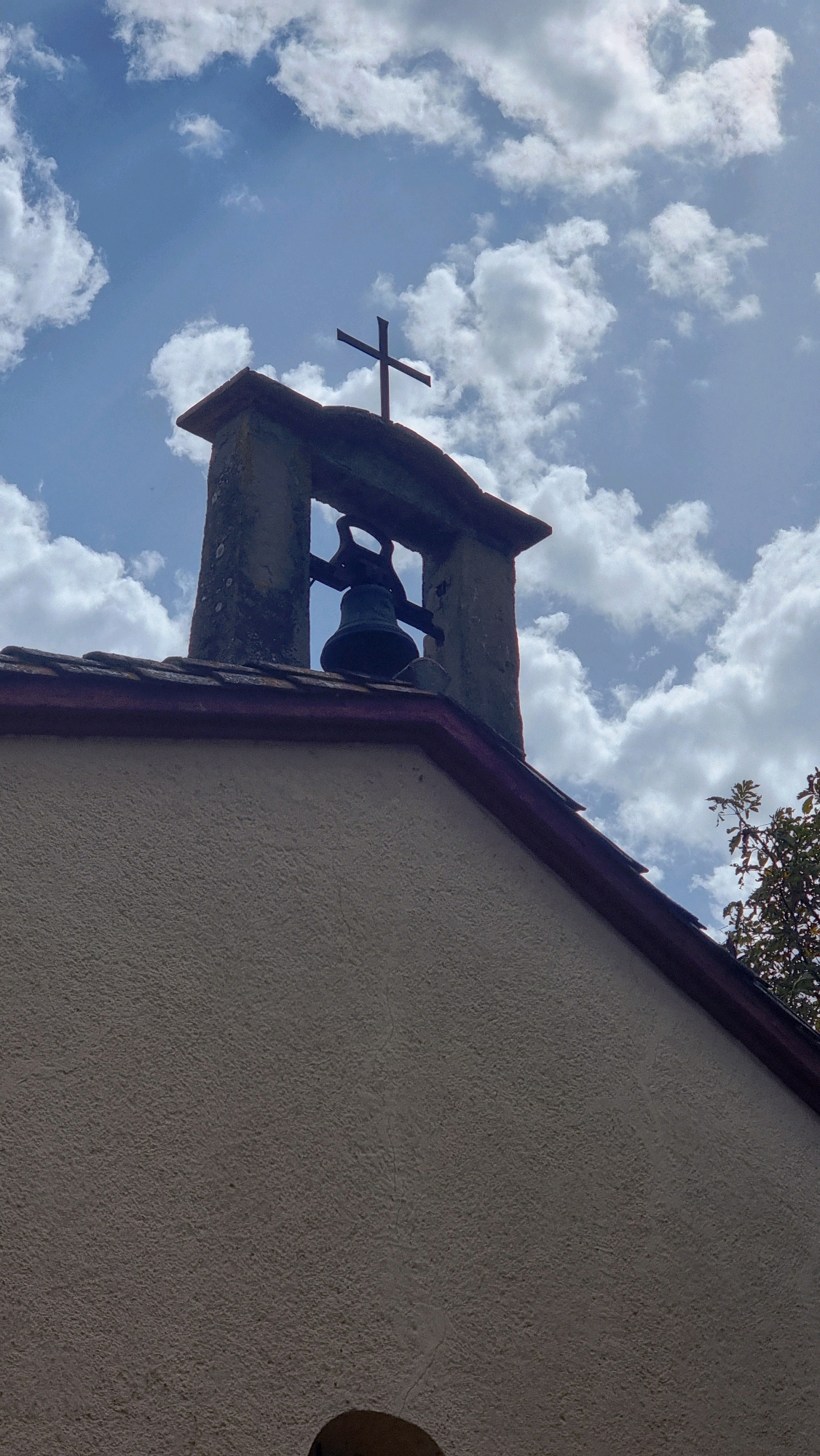 A low-angle, full shot captures a bell tower against a bright, partly cloudy sky. The tower, constructed of weathered stone, is topped by a simple cross. Below the cross, a dark bell hangs within the open frame of the tower. The roof of the building below the tower is visible, made of dark tiles with a red trim. The textured, cream-colored wall of the building occupies the foreground. The image appears to be taken on a sunny day in a rural or semi-rural setting, possibly in the town of la Vall de Bianya, Spain, given the architectural style. There are no people or animals visible in the frame.