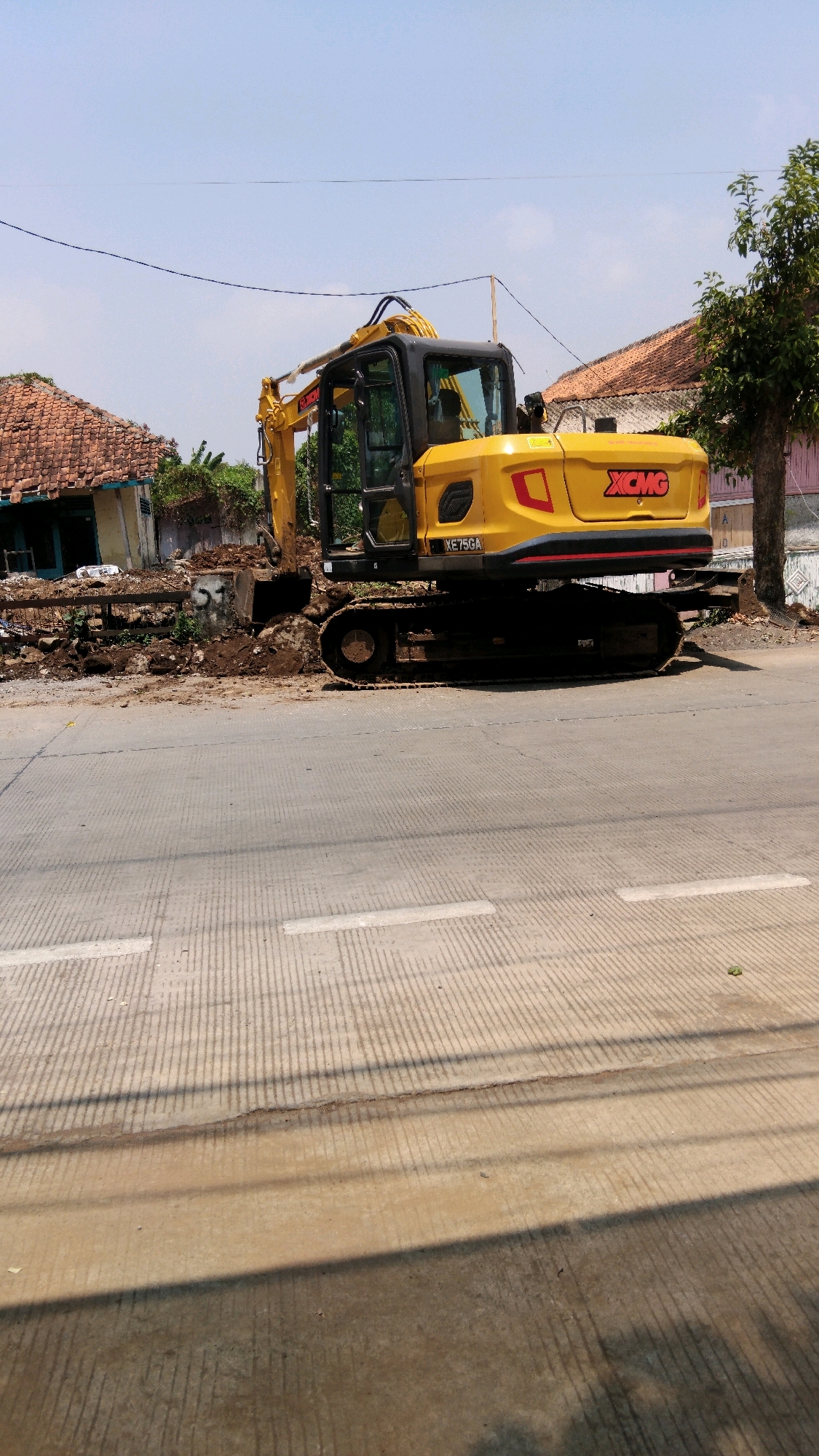 An outdoor, mid-angle shot captures a bright yellow XCMG XE75GA excavator parked on a dirt pile next to a paved road under a clear blue sky. The excavator's cab is visible, with a person seated inside. The machine's boom is angled upwards and slightly to the left, with the bucket resting in the dirt. To the left of the excavator, a dilapidated building with a damaged, tiled roof is situated. Beyond the building, lush green vegetation is visible. To the right of the excavator, a mature tree with green foliage stands. The road in the foreground is paved with a light gray concrete, featuring white lane markings. The scene appears to be in a semi-urban or rural area of Batang, Indonesia, possibly undergoing construction or demolition.