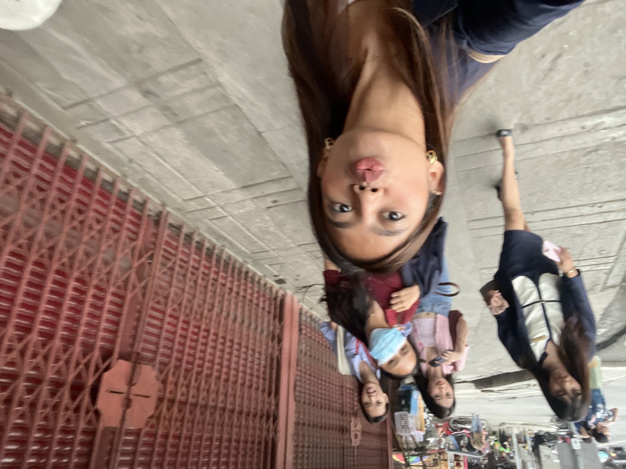 In Davao City, Philippines, a group of young women are walking on a sidewalk. The woman in the foreground is taking a selfie, puckering her lips. She is wearing a dark blue blazer and gold hoop earrings. Behind her, another woman in a red shirt and light blue face mask is visible, along with two other women. They are walking past a row of closed red metal gates. The environment suggests a bustling street scene, with some vehicles and shops in the background.