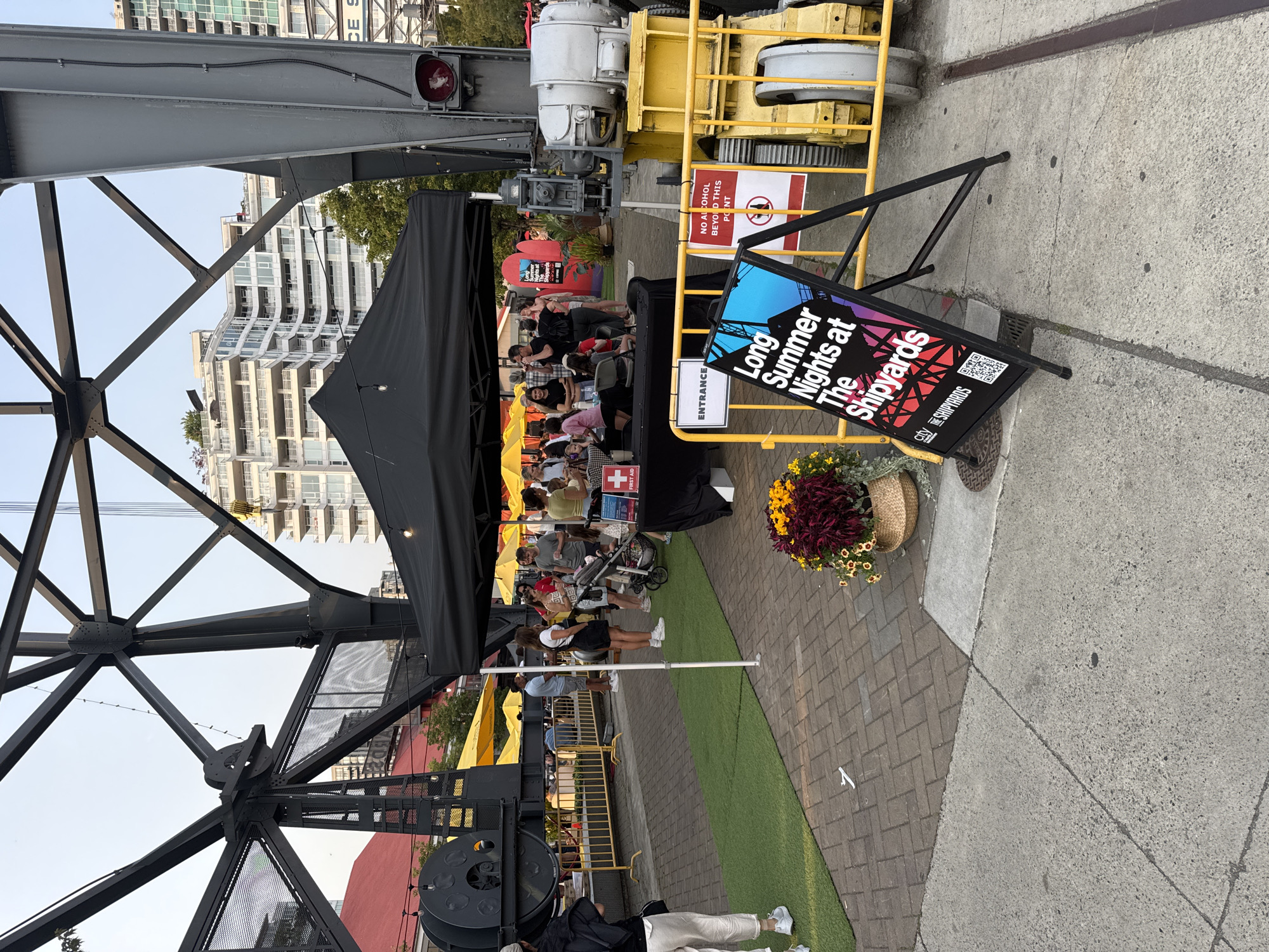 The image depicts an outdoor event at The Shipyards in North Vancouver, Canada. A black tent is set up with a black table displaying items, likely for sale or information. Several people are gathered around the tent and in the surrounding area, suggesting a public gathering or market. A prominent sign in the foreground reads "Long Summer Nights at The Shipyards" with a QR code. Yellow barriers are visible, separating different areas of the event space. The background features industrial structures, including a large black crane, and modern buildings, indicating an urban waterfront setting. Artificial turf is laid down on a section of the ground. A sign indicating "ENTRANCE" is placed on a yellow barrier. Another sign with a prohibition symbol and text "NO ALCOHOL BEYOND THIS POINT" is also visible. A decorative basket of flowers is placed near the sign.