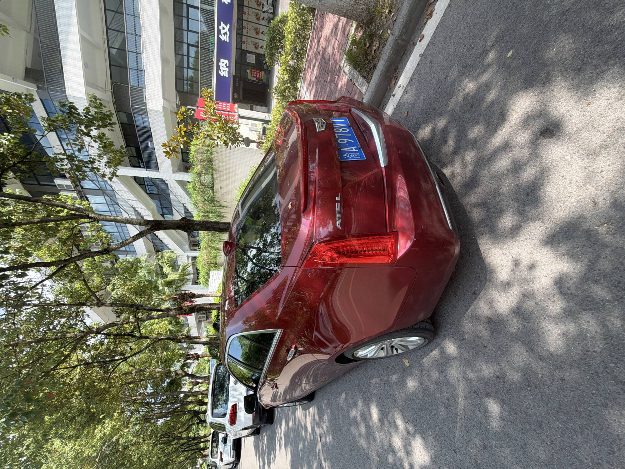 A red Cadillac ATS-L sedan is parked on the right side of an asphalt road in Jiangjin, China. The car is facing away from the camera, and its license plate reads "渝A 978V1". Behind the Cadillac, a white SUV is parked. A modern building with multiple levels and a glass facade is situated behind a row of trees lining the sidewalk. The sidewalk itself is paved with red brick. The scene is illuminated by sunlight, casting shadows from the trees onto the road.