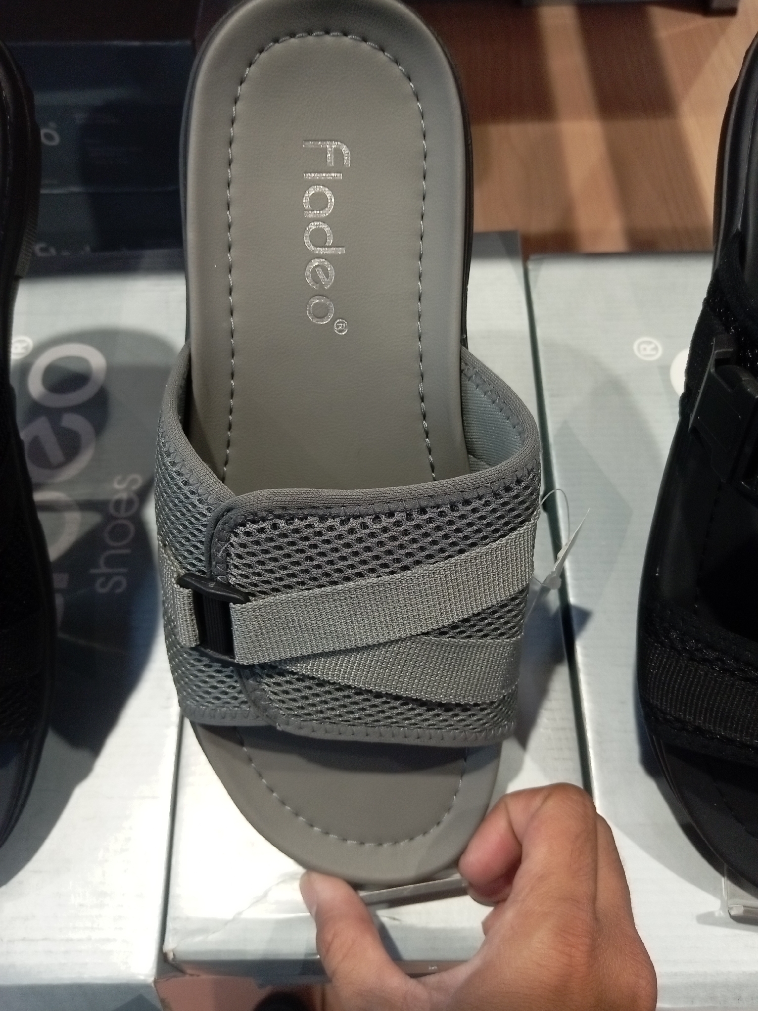 An overhead shot shows a pair of gray Fladeo brand sandals on display. The sandals are of a slip-on design with a single strap across the foot made of mesh and fabric. A black buckle is visible on the strap. The sandals are placed on top of white boxes. A human hand is visible at the bottom right of the frame, gently pressing on the sole of the sandal. In the background, another black sandal is partially visible. The scene appears to be a retail store environment, possibly a shoe store in Batam, Indonesia, given the text visible on the boxes in the background, which appears to be "Batam Shoes".