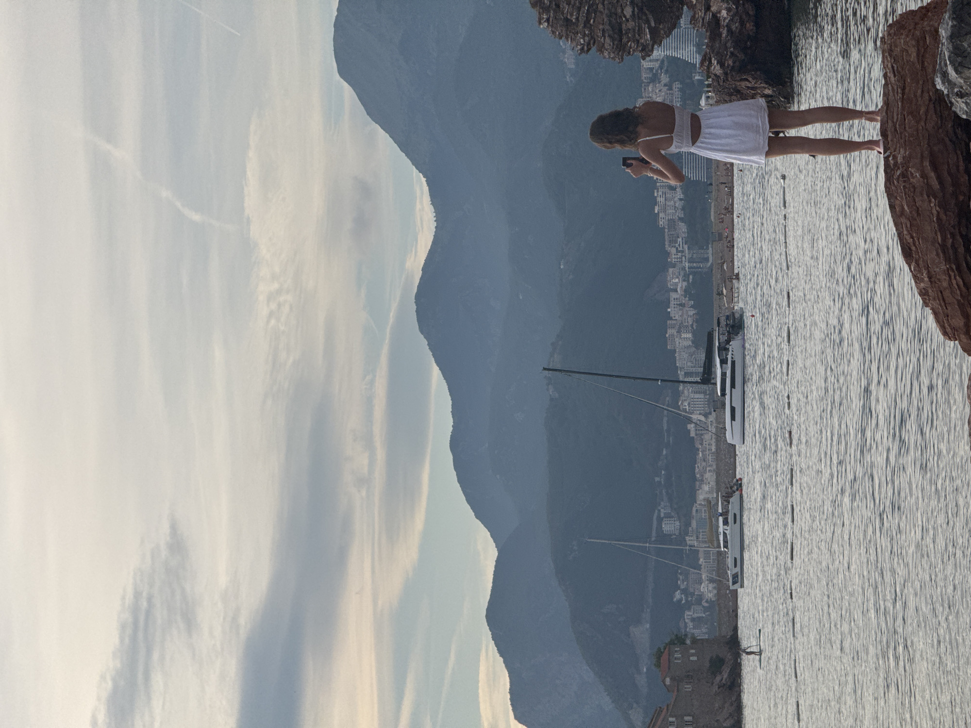 In Kamenovo, Montenegro, a woman stands on a large rock, facing away from the viewer, holding a smartphone to take a photograph. She is wearing a white dress and her feet are bare. The sea is calm and reflects the sky, which is partly cloudy with a few contrails from airplanes. In the background, mountains rise behind a coastal city with a mix of modern and older buildings. Several sailboats are docked in the harbor. To the left, a person on a paddleboard is visible near a stone structure. The overall scene depicts a serene coastal view with a solitary figure capturing the moment.