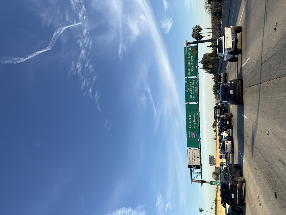 The image shows a congested highway scene under a clear blue sky with wispy clouds. Traffic is bumper-to-bumper, with a mix of cars and trucks visible. Overhead green highway signs indicate directions: South to LAX Airport and Long Beach via the 405, Slauson Avenue/Marina del Rey via the 90, and Washington/Culver Boulevards at the next exit. Additional signs point out the carpool lane.