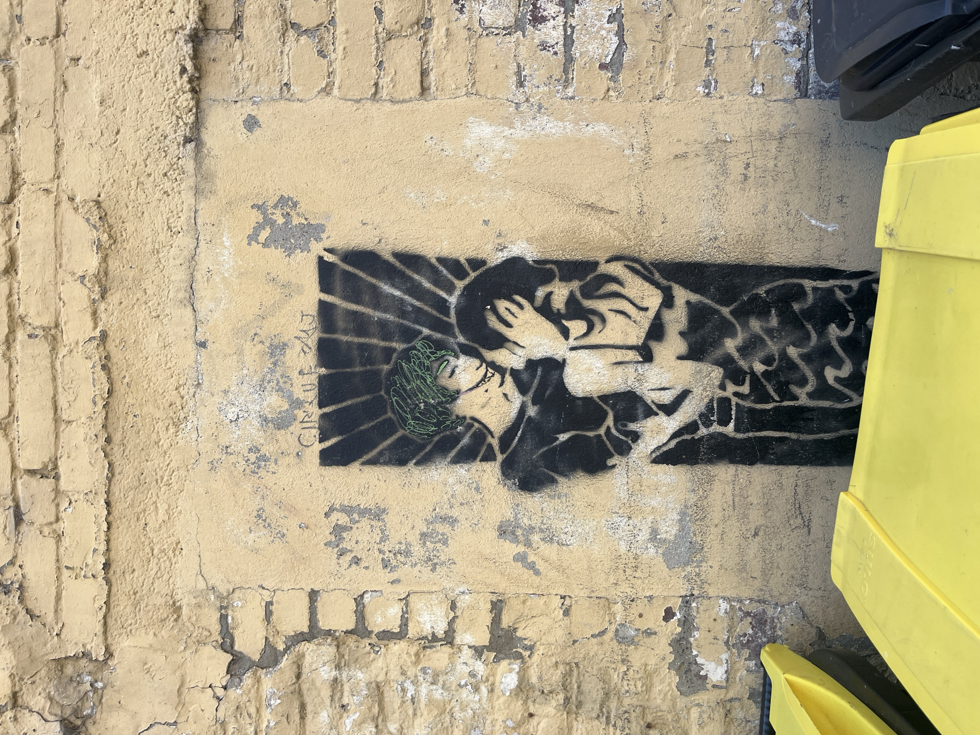The image shows a stencil graffiti of a couple embracing and kissing on a weathered yellow brick wall. The graffiti is in black and white with green accents on the hair of one of the figures. The wall exhibits signs of age and damage, with peeling paint and crumbling sections. In the foreground, two bright yellow trash bins are partially visible, along with a portion of a dark grey trash bin. The graffiti is centered on the wall, with a dark, sunburst-like pattern behind the couple. The text "CIRQUE DU J" is faintly visible above the graffiti. This artwork is located on a street in Dieppe, France.