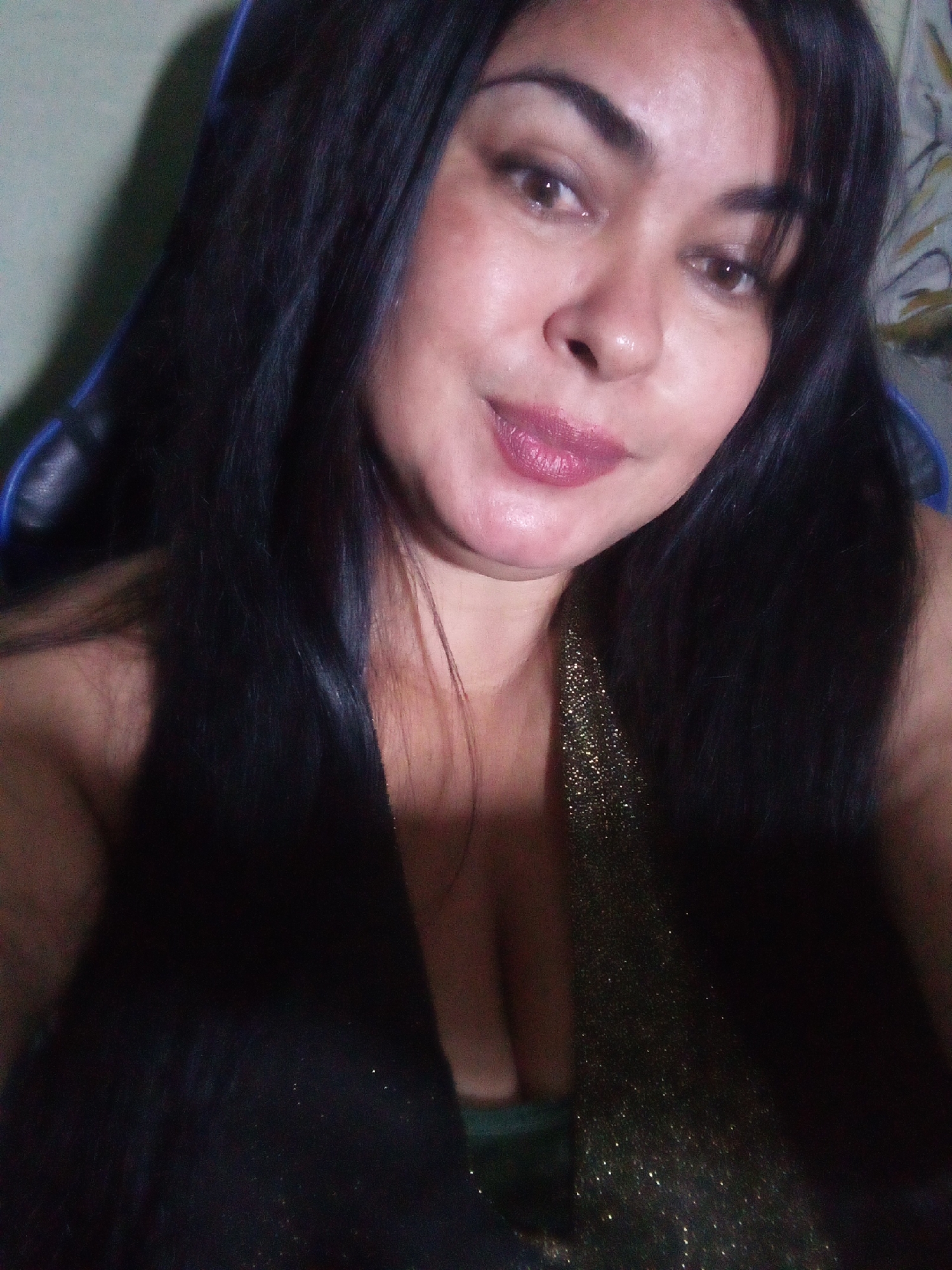 A woman with long, black hair and tan skin is facing the camera. She has dark eyebrows and eyes, and her lips are a mauve color. She appears to be wearing a black top, with a section of gold, sparkly fabric also visible. The background is blurry and indistinct, but it includes a light green wall and what appears to be part of a blue chair. There is no visual information in the image to support identifying Juan Griego, Venezuela as the location.