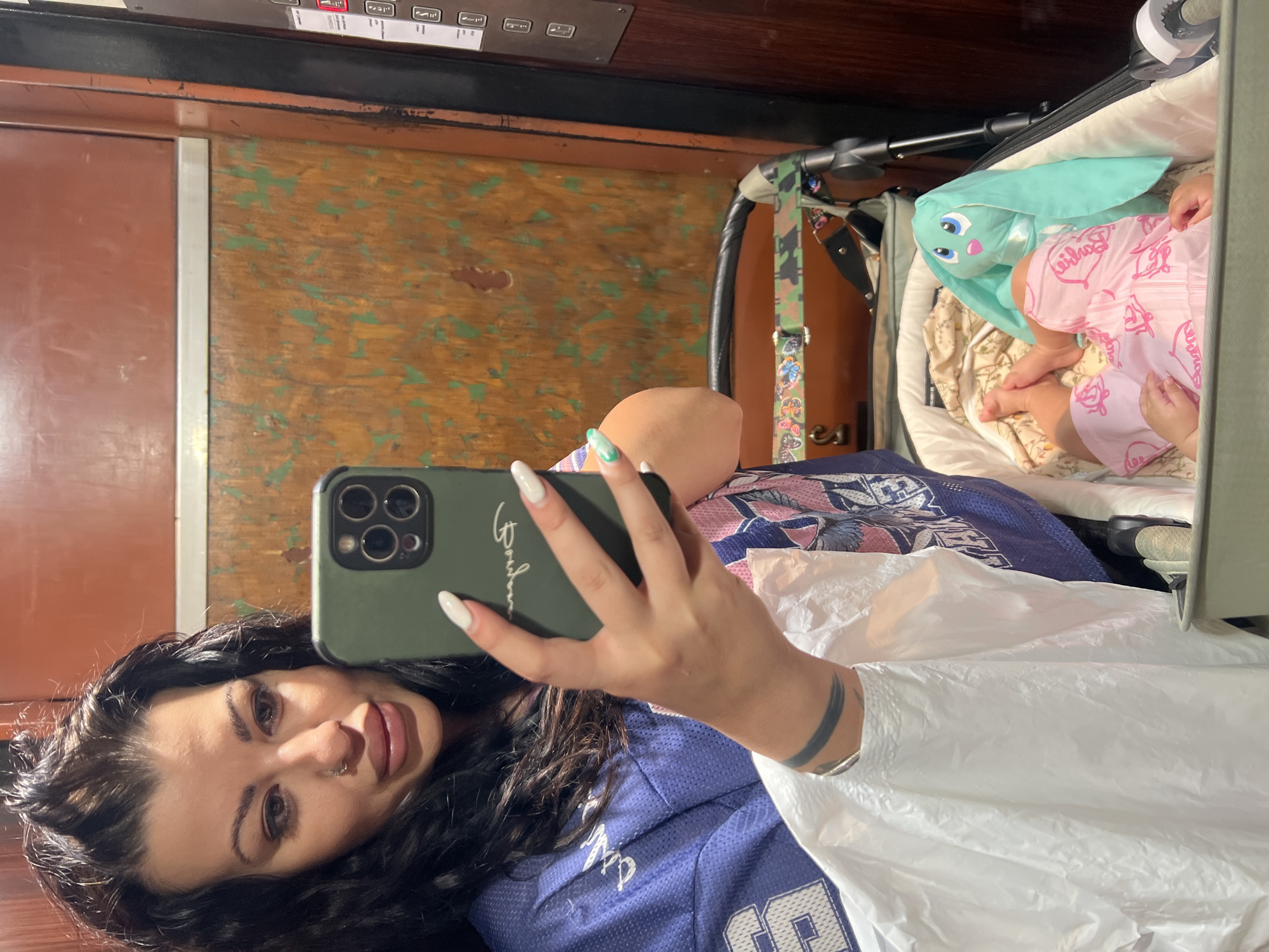 A young woman takes a selfie in what appears to be an elevator with a brown wooden interior. She is holding a smartphone with a green case, her face is visible in the photo, she has dark hair and multiple piercings. She is wearing a blue jersey, and what looks like a disposable poncho. In the background, there is a baby in a stroller with a mint green rabbit stuffed animal next to them. The exact location in Plovdiv, Bulgaria, cannot be determined from the provided image.
