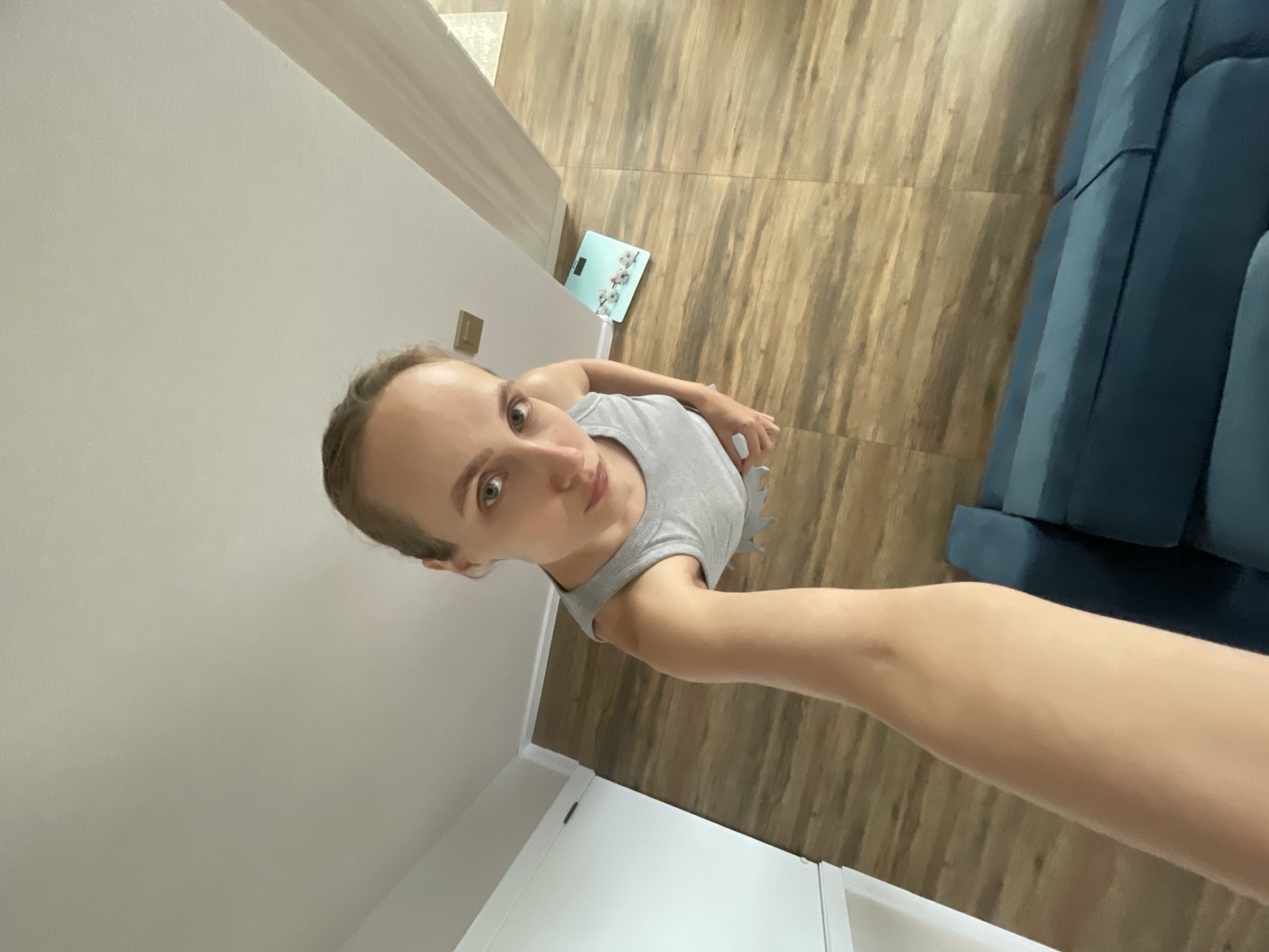 A young woman is taking a selfie in a room. She is wearing a grey tank top. The room has light-colored walls and a wooden floor. In the background, a teal bathroom scale with a floral design is visible. A dark blue couch is partially visible on the right side of the image. The image appears to be taken indoors, during the daytime. The woman's expression is neutral. There is no visible text in the image.