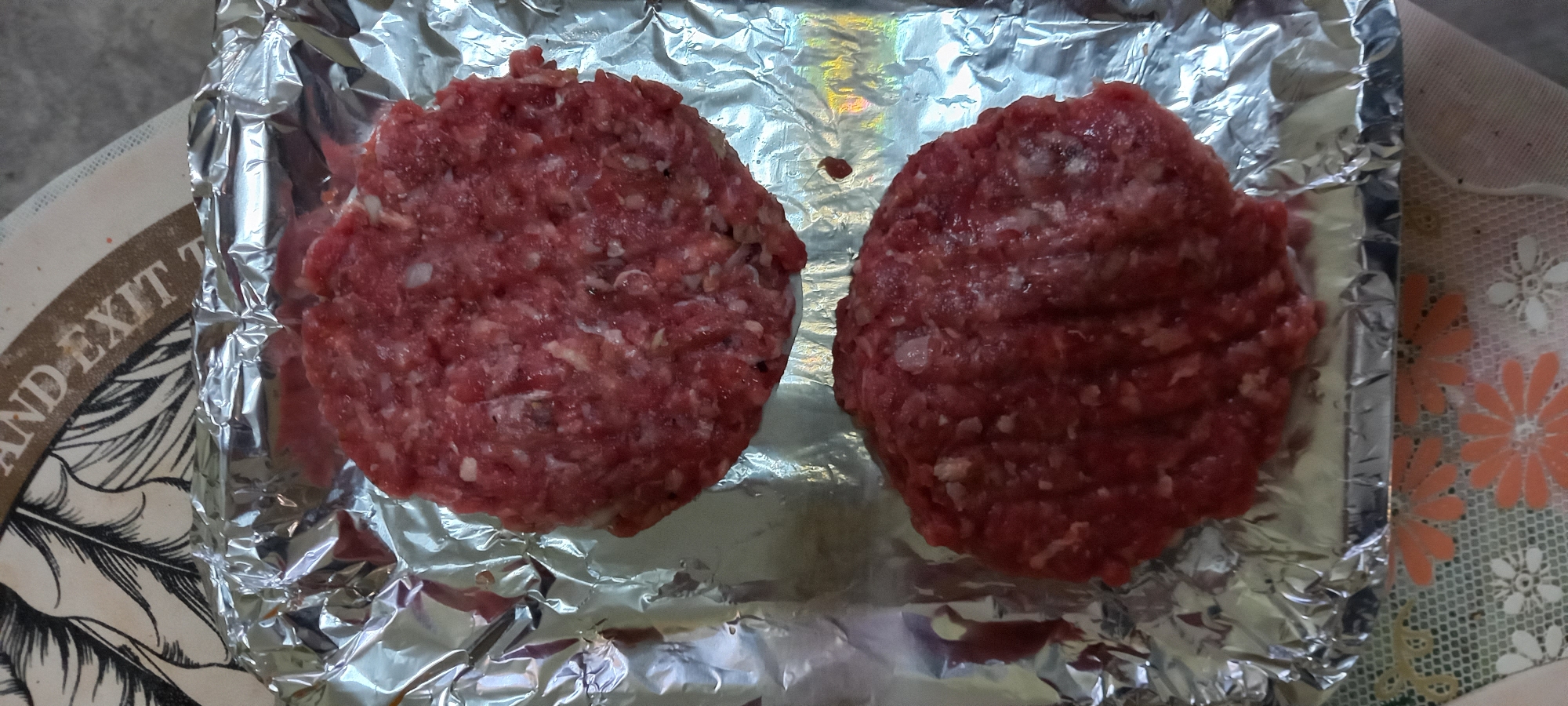 The image shows two raw hamburger patties on a sheet of aluminum foil. The patties are round, with a coarse texture of ground meat and small flecks of white, presumably fat. The aluminum foil is crumpled and reflective, showing various light sources in its surface. In the background, partially visible around the foil, are two different surfaces: a light-colored, possibly cream-colored, surface with a pattern that includes black feathers and the words "AND EXIT T" and a lacy surface with an orange flower pattern. The photograph appears to have been taken indoors. The city of Baan Chaloklum, Thailand is not explicitly identifiable within the image content.