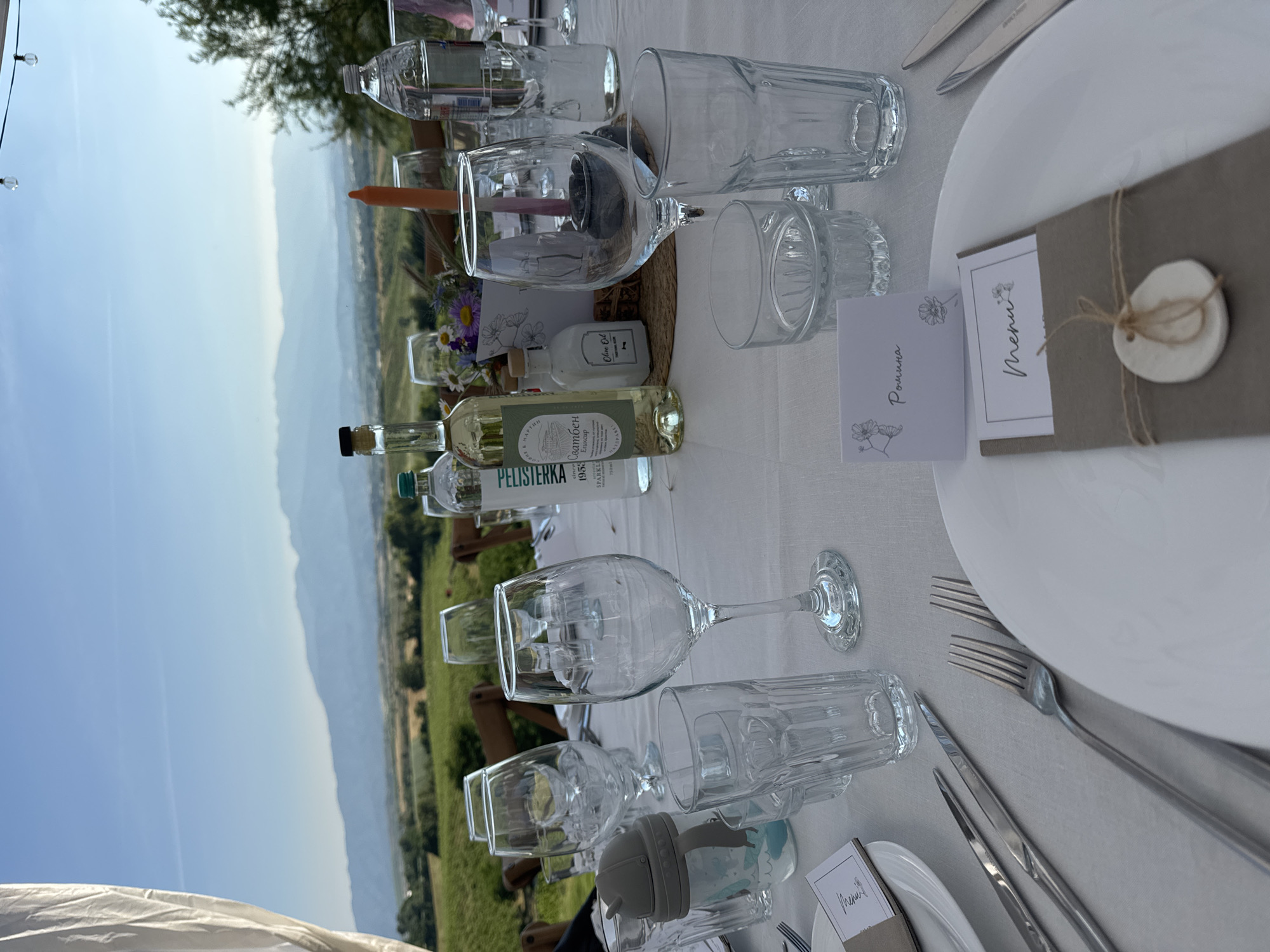 This image captures an outdoor table setting prepared for an event or meal, with a panoramic view of the natural landscape near Harsovo, Bulgaria. The scene is illuminated by clear daylight under a bright blue sky.
In the foreground, a rectangular table is covered with a crisp white tablecloth. Multiple place settings are arranged, featuring white ceramic plates, polished silver forks, and knives. A variety of glassware is present, including stemmed wine glasses, cylindrical tumblers, and smaller drinking glasses. A gray baby sippy cup with a blue design is also positioned on the table.
Individualized place cards and menus are visible. One prominent place card in the lower foreground reads "Ромина" (Romina) in cursive script, adorned with line-art floral motifs. Adjacent to it, a menu card is nestled within a brown fabric sleeve, tied with a natural twine bow and a small, white, circular ceramic embellishment.
Along the table's center, decorative elements include a small vase holding a mixed bouquet of wildflowers (daisies and purple blossoms), a tall orange taper candle, a small white bottle labeled "Olive Oil," a clear bottle of "PELISTERKA SPARKLE 1953" mineral water, and a white wine bottle with a label displaying "Сватбен Еликсир" (Wedding Elixir). Some of these items rest on a circular woven placemat.
The background unfolds into a expansive vista of rolling green hills, cultivated fields, and distinct rows characteristic of vineyards, extending towards a distant range of hazy blue mountains that demarcate the horizon beneath a clear blue sky. A white fabric drape or canopy is partially visible on the far left side of the frame, and a string of outdoor lights with visible bulbs hangs above the table in the upper right portion of the image. There are no individuals depicted in the scene.