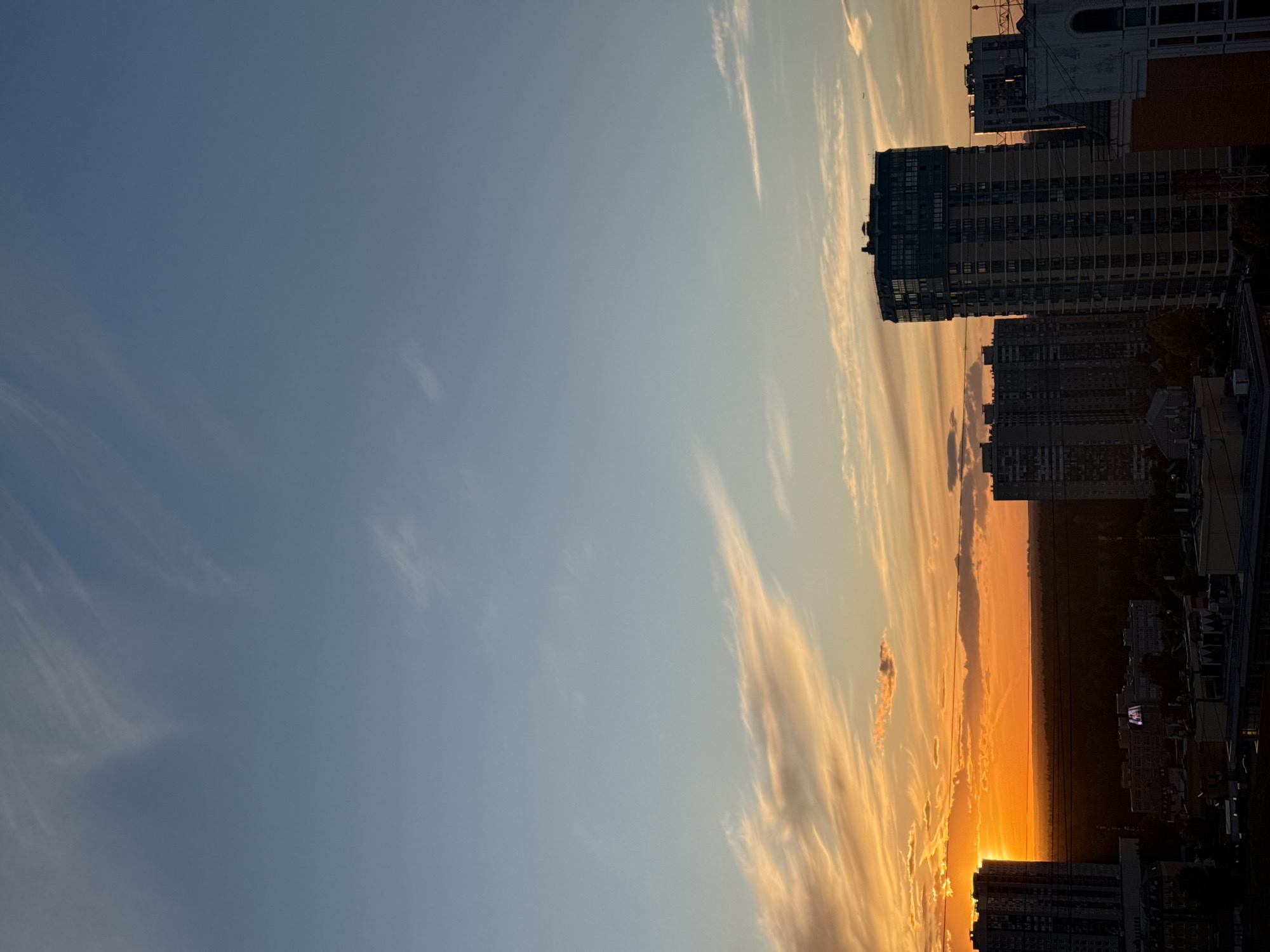 This is a photograph taken in Khimki, Russia, depicting a sunset over an urban landscape. The sky is a gradient of soft blues and oranges, with wispy clouds stretched across it. The setting sun is visible on the horizon, casting a warm glow over the scene. Several tall buildings, characteristic of a city environment, are silhouetted against the vibrant sky. One prominent building, with a dark facade and many windows, dominates the right side of the frame. The overall mood is serene and peaceful, typical of a sunset. No people or specific activities are visible. The image appears to be a wide shot, capturing the expansive sky and the urban sprawl below.