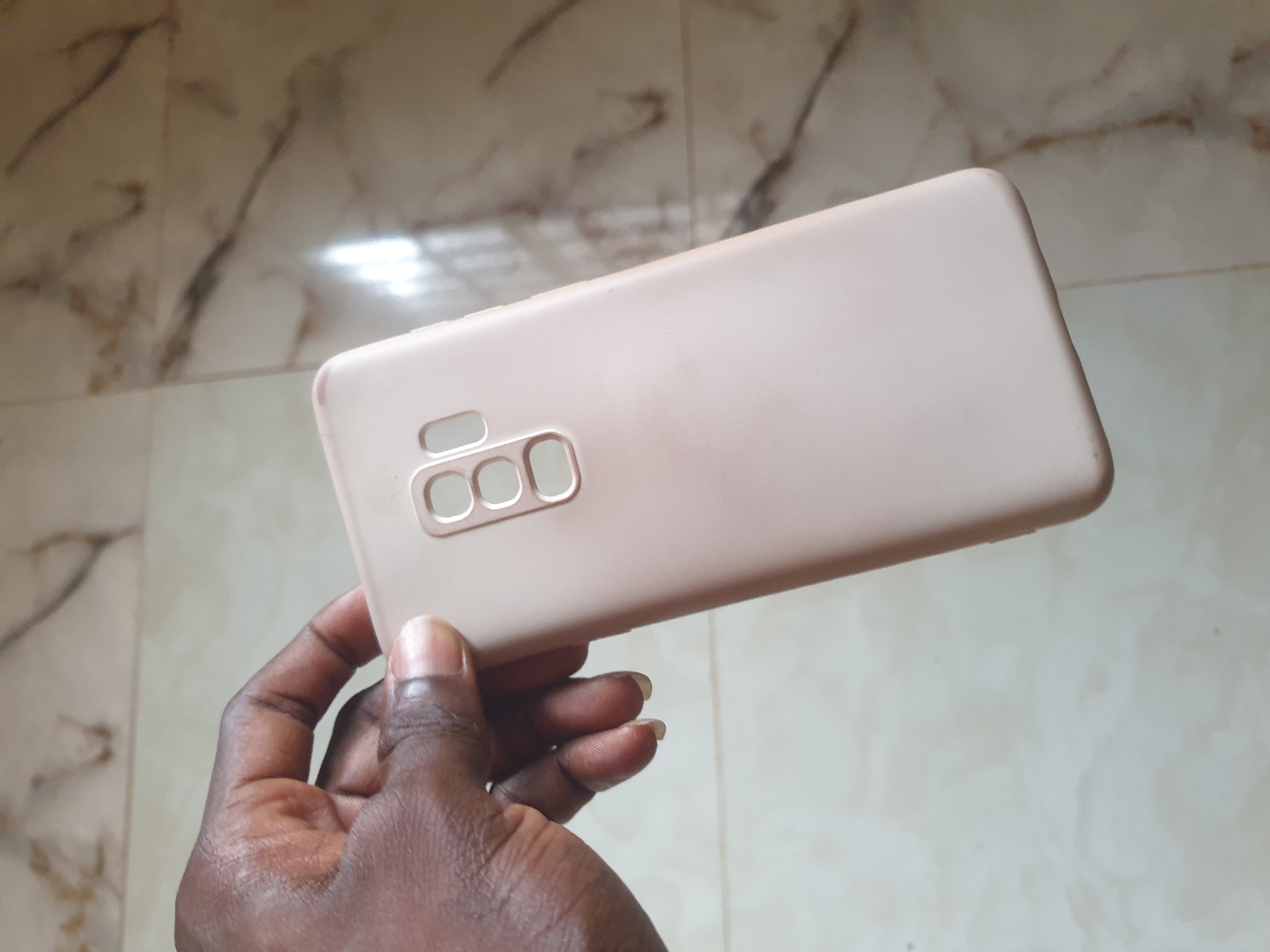 A hand holds a light pink phone case with three cutouts for a camera lens array. The phone case is positioned in the center of the image, with the hand gripping the left side. The background consists of a tiled floor with a marble-like pattern. The lighting appears to be natural, casting subtle shadows.