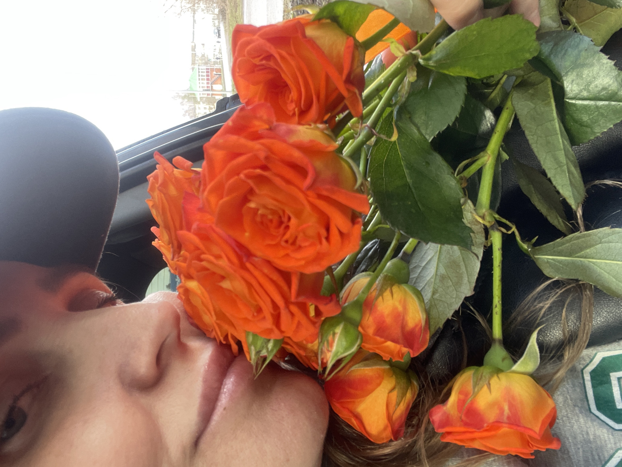 A close-up shot features a person's face partially visible, seemingly within a vehicle. The individual is wearing a black cap. The focus is on the person's face, which is close to a bouquet of orange roses with green leaves and stems. The background shows a blurred cityscape, likely a residential area with buildings visible through the vehicle's window. While Chaikovsky, Russia, is mentioned, there is no clear indication from the image that it is taken in that location.