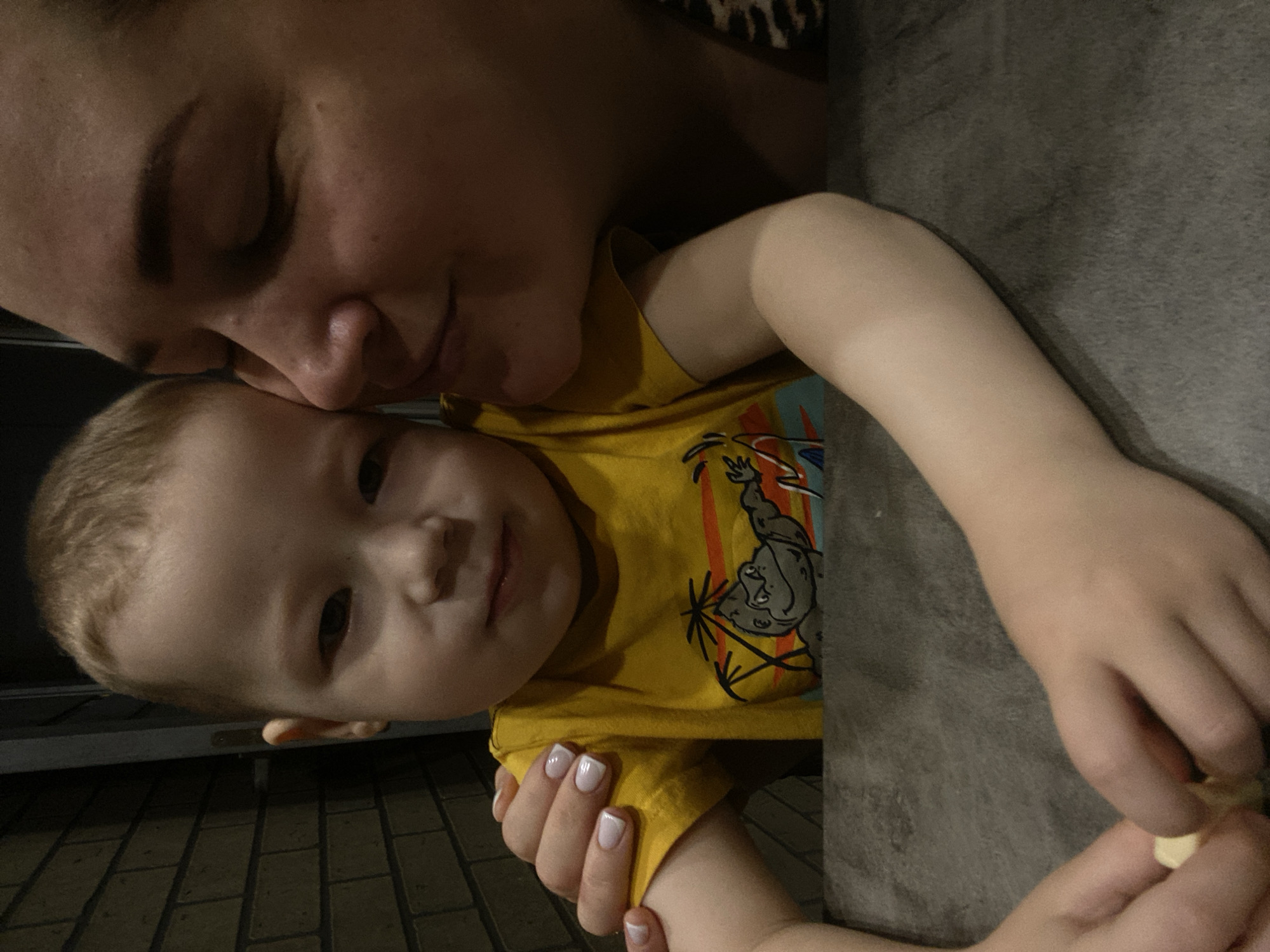 A woman and a young child are in an indoor setting, possibly a restaurant or café. The woman is positioned at the top of the frame, her face close to the child's. The child is wearing a yellow shirt with a cartoon graphic. One of the child's arms rests on a grey surface. The woman has manicured nails. The child's other arm is held in the woman's hand. In the lower-left corner, a dark, patterned floor is visible.
