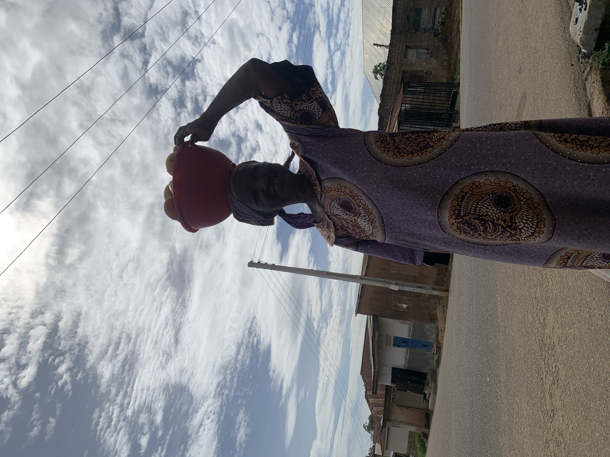 An older woman of dark complexion stands on an asphalt road in Jos, Nigeria, facing the viewer with a slight smile. She wears a long, loose-fitting purple garment adorned with large, circular brown, gold, and white patterns, complemented by a matching headscarf. On her head, she balances a red plastic basin containing several light-colored, spherical objects, presumably fruits, steadying it with her raised left hand.
The environment consists of a paved, granular asphalt road in the foreground and midground. In the background, a cluster of single-story buildings with various wall finishes (some plastered light gray, some unplastered earthen tones) and corrugated metal roofs (some rusty, some metallic silver) line the road. One building on the left features a blue door and a white sign with black text, partially legible as "FEDERAL GOVERNMENT OF NIGERIA" and "MINISTRY OF". A concrete utility pole with multiple overhead power lines is visible behind the woman to her left. Additional power lines traverse the upper sky from left-center to upper right. The sky is partly cloudy with patches of bright light suggesting diffused sunlight, and numerous white stratocumulus clouds are scattered across the blue sky. A dark metal gate is visible in the background on the right, near another building with a prominent silver corrugated roof.