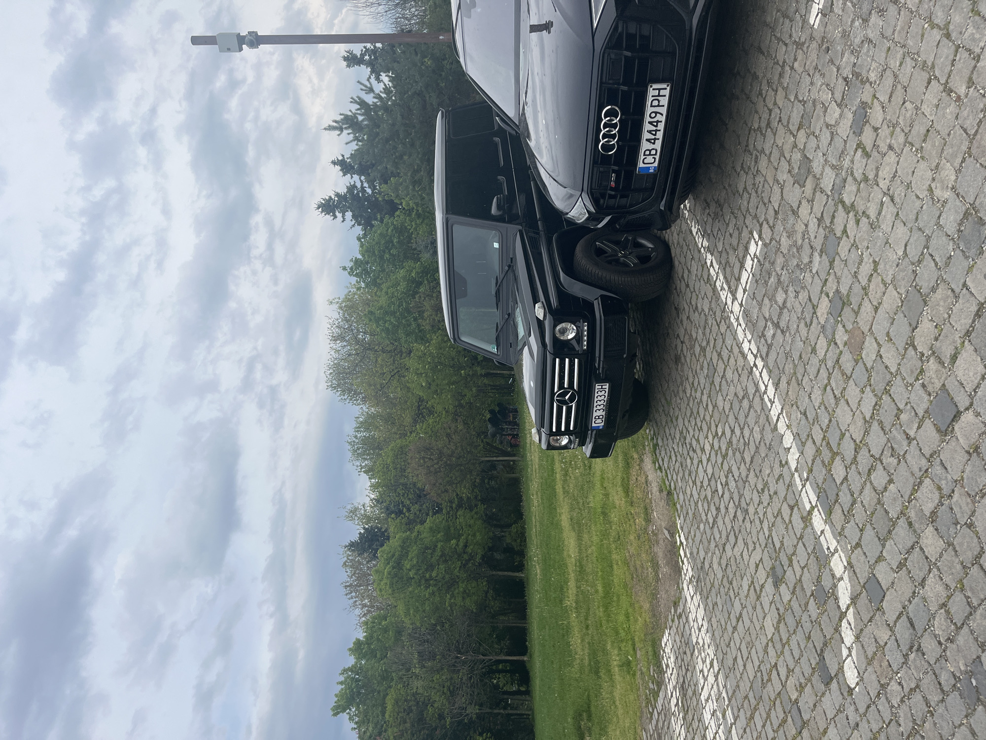 This rotated daytime image captures two black luxury SUVs parked closely side-by-side on a cobblestone surface, marked with white parking lines. Adjacent to the parking area is a lush green grassy field, backed by a dense line of tall green trees under a predominantly overcast sky with scattered clouds.
The vehicle on the left (when oriented correctly) is a Mercedes-Benz G-Class, facing forward. Its distinctive vertical slat grille, circular headlights, and the Mercedes-Benz emblem are clearly visible. Its Bulgarian license plate reads "CB 3333H".
The vehicle on the right (when oriented correctly) is an Audi SUV, specifically an SQ model based on the "S" badge and aggressive grille design. The prominent Audi rings logo is centered on its large black grille. Its Bulgarian license plate, "CB 4449 PH", is affixed to the front bumper.
In the upper portion of the image, against the cloudy sky, a tall, slender utility pole can be seen, fitted with a white rectangular device or sensor near its top. The "CB" prefix on both license plates identifies the vehicles as registered in Sofia, Bulgaria, consistent with the provided location context. No people are visible, and the scene depicts a quiet outdoor parking area.