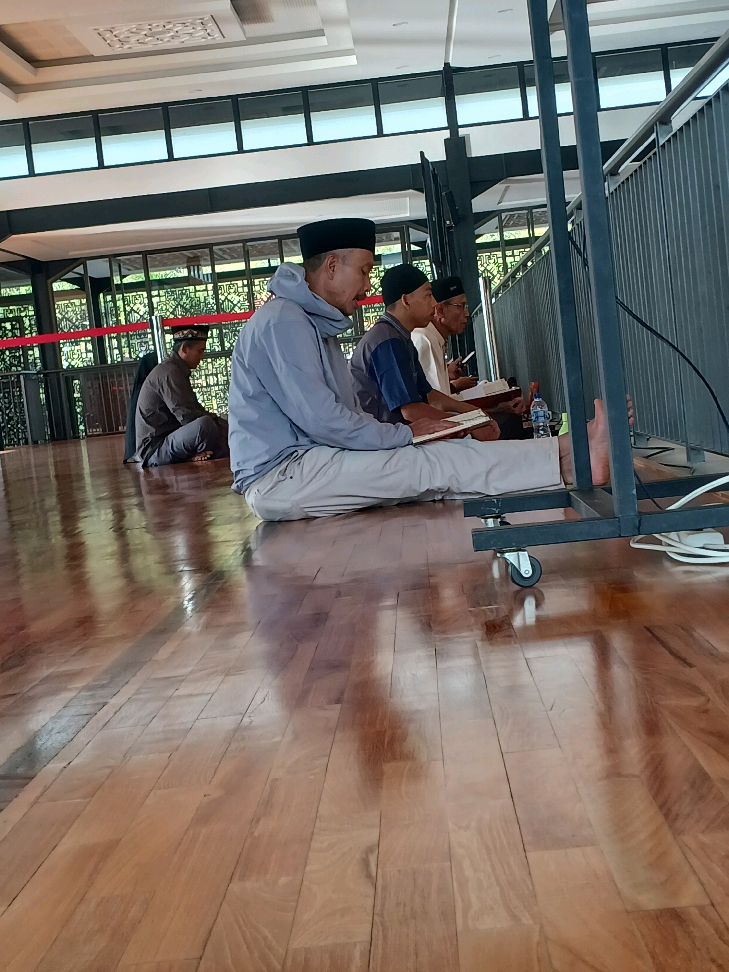 Four men are seated on a polished wooden floor in what appears to be a covered outdoor space. The man in the foreground on the left is wearing a light blue hooded jacket and light-colored pants, with a black skullcap. He is looking down at a book or papers in his lap. To his right, another man in a dark blue shirt and black skullcap is also looking down at a book. Further to the right, a third man in a light-colored shirt and glasses is looking down at a book, and a fourth man in a dark brown garment and a patterned cap is seated to the far left, facing away from the camera. The space has a modern ceiling with decorative panels and large windows looking out onto greenery. Red barriers are visible in the background. A metal structure with wheels, possibly a stand, is in the right foreground. The floor is highly reflective, showing the ceiling and overhead lights. The scene suggests a gathering for religious study or prayer. The location is identified as Arjasari, Indonesia.