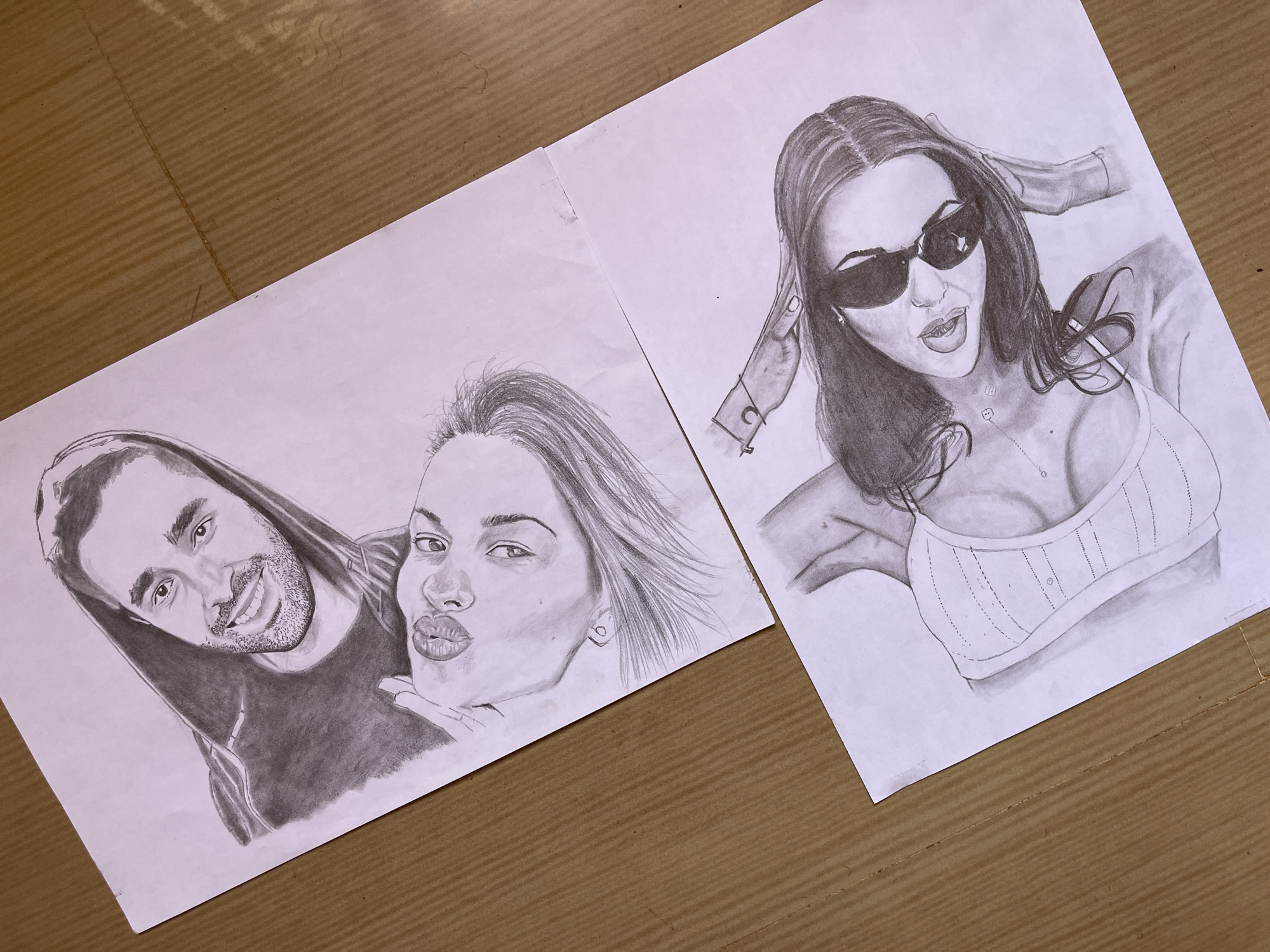 Two pencil drawings are laid out on a brown, ridged surface.
The drawing on the left depicts a man and a woman posing closely together. The man, on the left, has short dark hair, a beard, and is smiling. He wears a dark hooded sweatshirt. The woman, on the right, is pouting her lips in a kissing expression. Her hair is windswept.
The drawing on the right depicts a woman wearing sunglasses and a low-cut tank top. Her hair is dark, and she has a necklace. She is holding her hand near her head.
The location of the city of Jos, Nigeria is not discernible in this image.