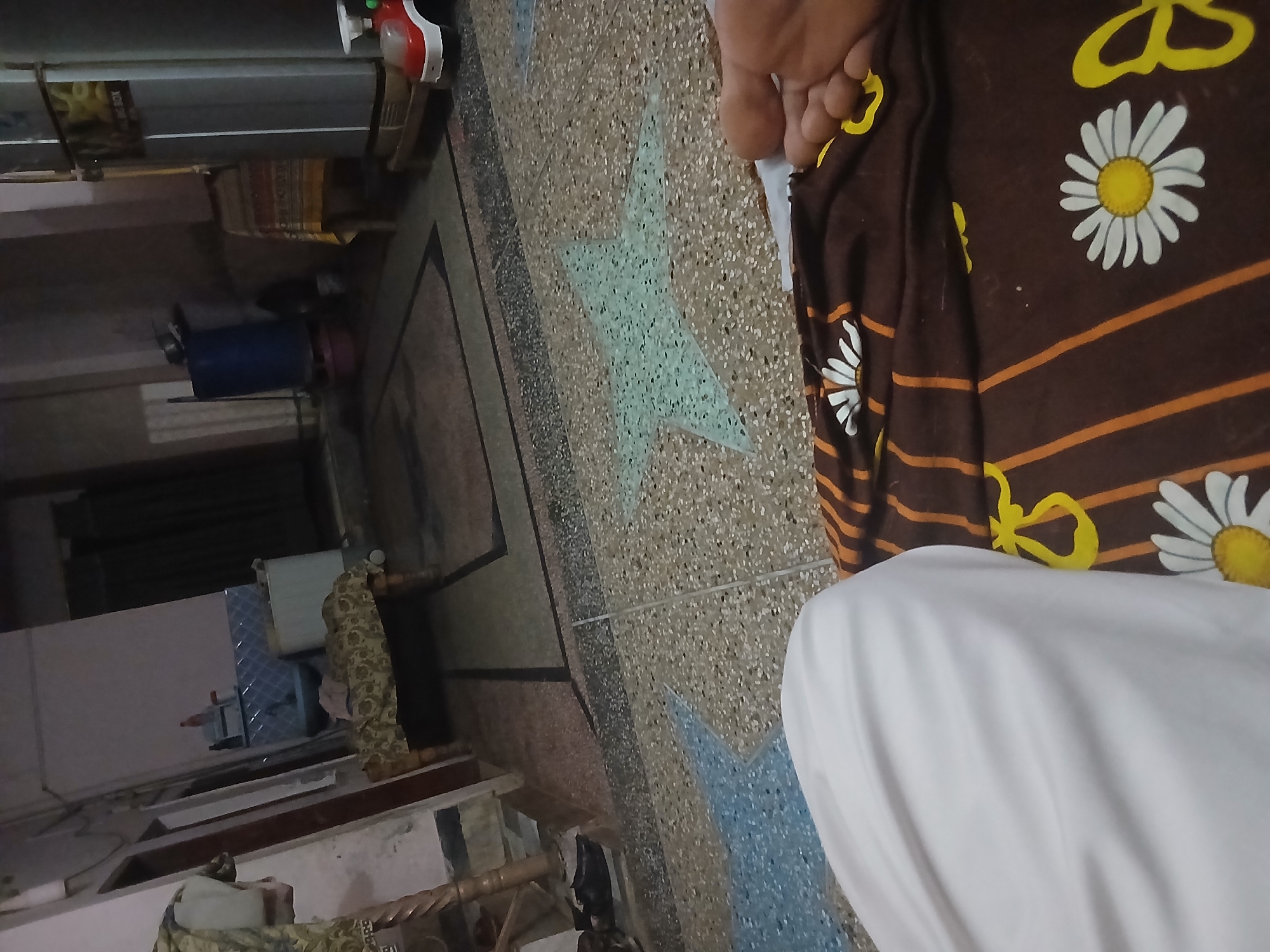 The image depicts an indoor scene, likely a residential space, in Peshawar City Tehsil, Pakistan. In the foreground, a person's bare foot is visible, resting on a brown blanket with a daisy pattern. To the left, a white cloth is draped, obscuring part of the person. The floor has a speckled pattern with large star shapes. In the background, a refrigerator is visible on the right, with a small red toy car in front of it. A blue barrel and a table covered with a patterned cloth are also present further back. The overall environment appears somewhat cluttered and suggests a lived-in space.
