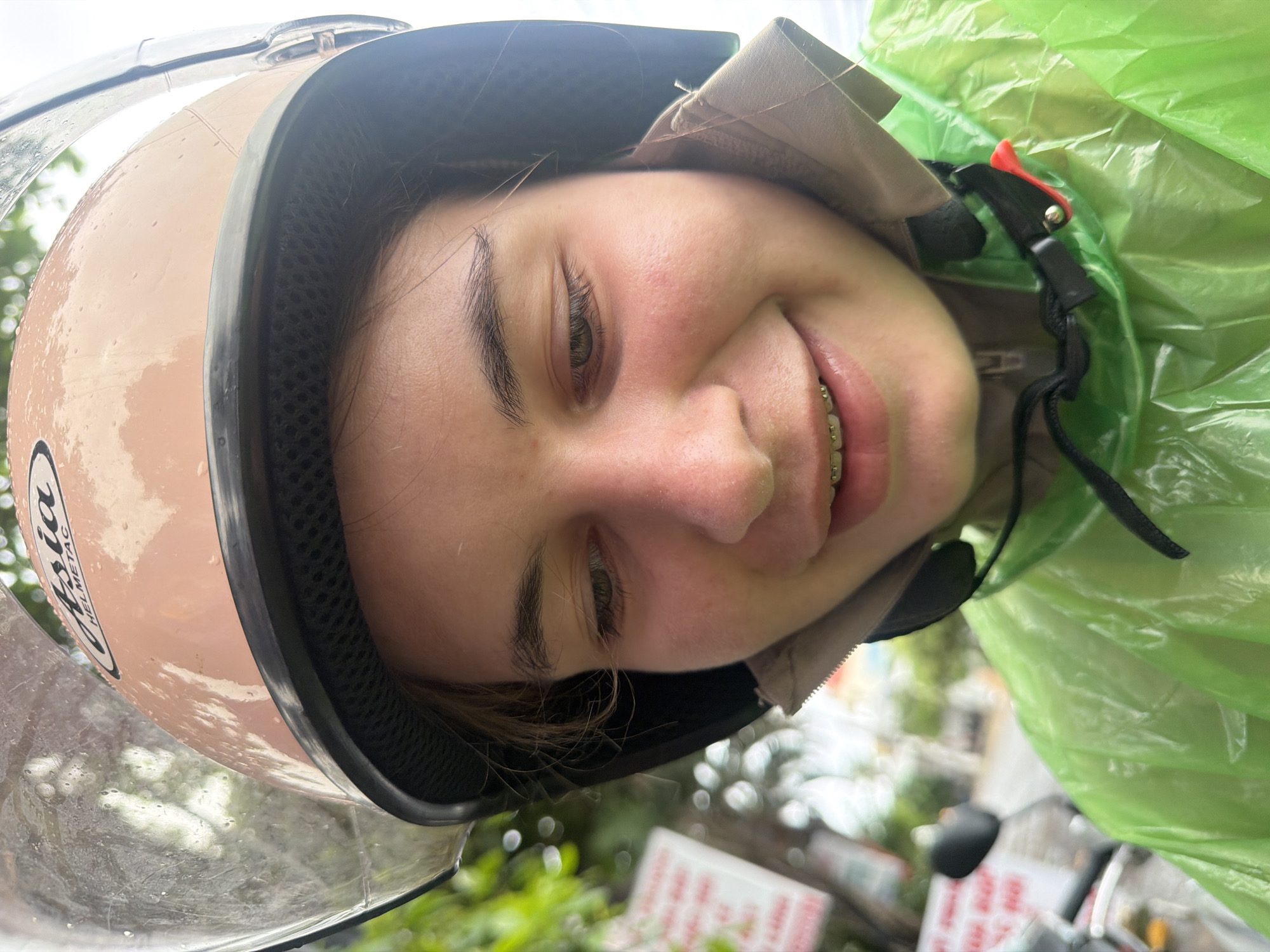 A close-up, waist-up shot features a young person wearing a light pink motorcycle helmet with a clear visor. The helmet has a black lining and the brand "Asia Helmetac" is visible on the top. The person is also wearing a bright green, translucent raincoat over a beige jacket. Braces are visible on the person's teeth as they smile. The background is blurred, showing hints of greenery and possibly street signs, suggesting an outdoor urban setting in Nha Trang, Vietnam. The lighting is natural, and the image appears to have been taken during the daytime.