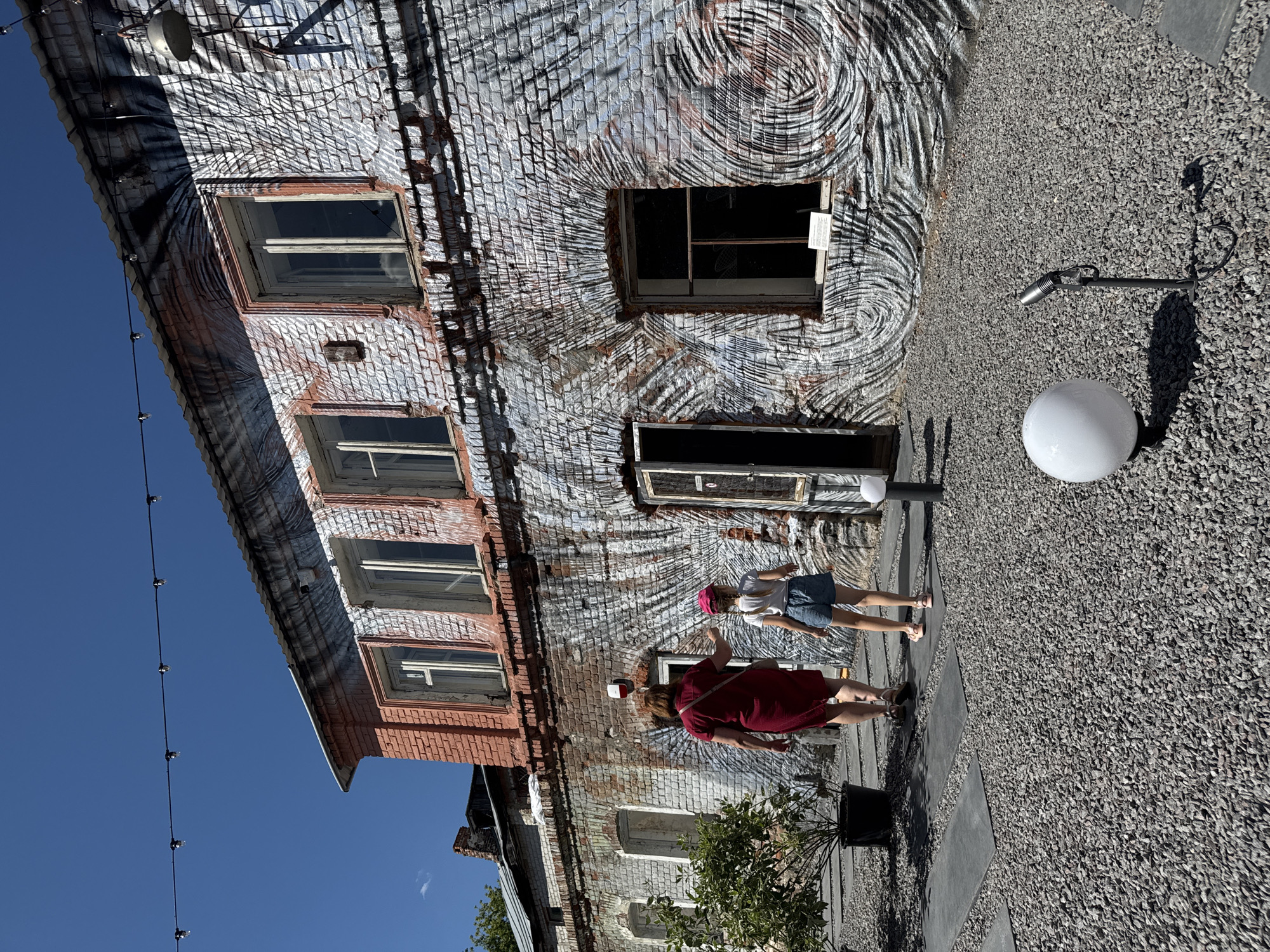 In Yelets, Russia, an adult woman in a red dress and a young girl wearing a pink hat and blue shorts walk on a gravel path towards a brick building. The building features a distinctive mural on its exterior walls, with a pattern resembling concentric circles and brush strokes. The mural is painted over aged bricks, some of which are red and others are a lighter, weathered hue. The building has multiple windows, some of which are open. To the left of the building, a potted plant sits near the path. The ground is covered in dark gray gravel, interspersed with dark gray rectangular stepping stones. In the foreground, a white globe-shaped lamp stands on a short black pole, and a small, dark spotlight on a stand is also visible. Overhead, a string of bare light bulbs hangs against a clear blue sky. The scene appears to be an outdoor courtyard area, possibly a public space or a commercial establishment, given the art on the building and the decorative lighting.