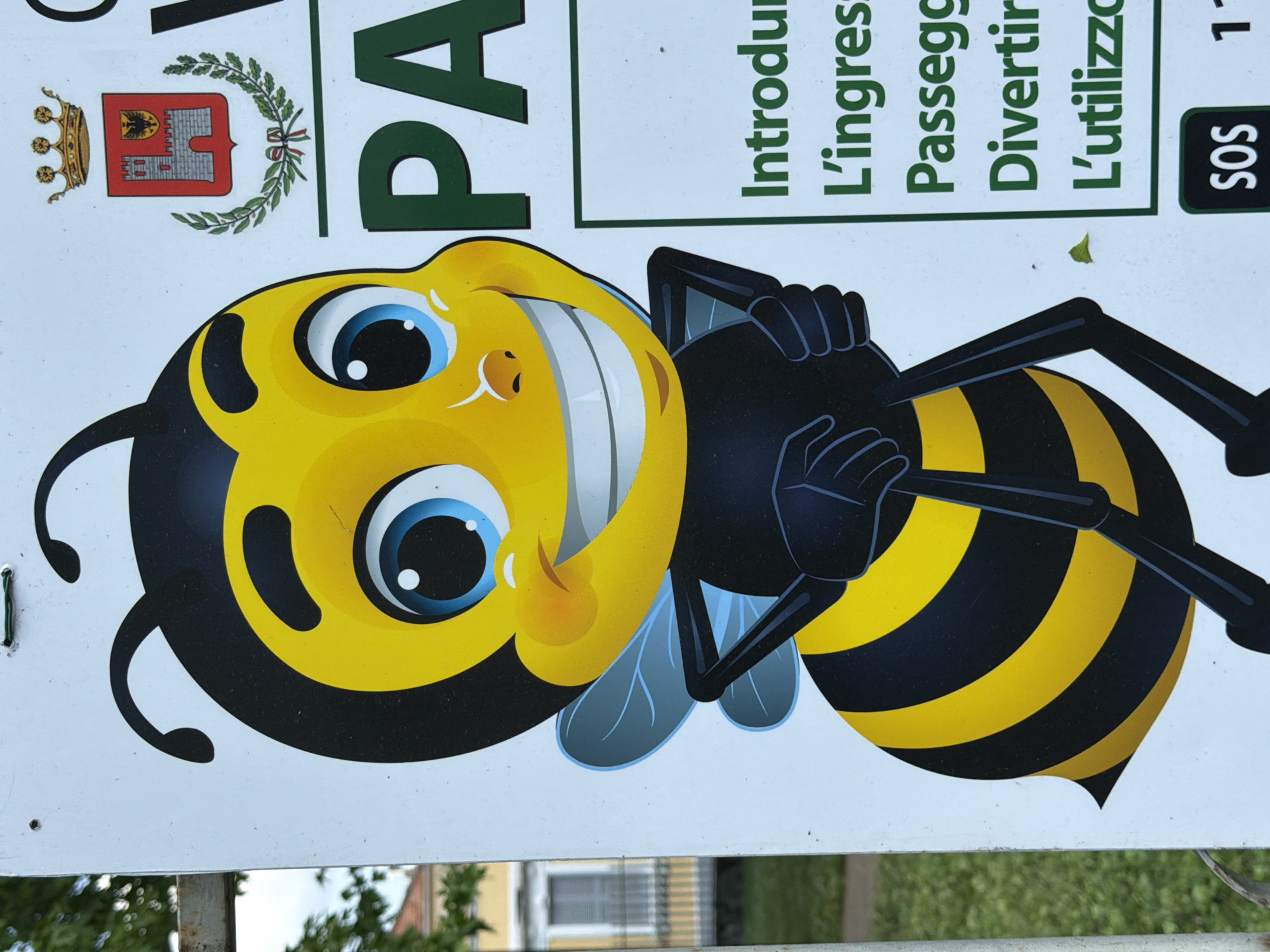 The image captures a portion of a white outdoor sign, likely located in a public area in Vigevano, Italy, given the municipal crest. The sign is dominated by a large, cheerful cartoon bee character with a black and yellow striped body, light blue wings, and antennae. The bee has a wide, happy smile with visible white teeth and large, expressive blue eyes, conveying a friendly and welcoming demeanor.
In the upper part of the sign, an official-looking crest is visible. It features a red shield with a grey castle-like structure, topped by a golden crown, and framed by a green wreath of laurel and oak leaves, characteristic of a municipal coat of arms.
Several lines of Italian text are present. Partial words like "PA" are visible, likely part of a larger word such as "PARCO" (Park) or "PASSEGGIATE" (Walks). Other clear words include "Introdu" (possibly from "Introduzione" or "Introdurre"), "L'ingress" (L'ingresso - The entrance), "Passegg" (Passeggiate - Walks), "Divertir" (Divertimento - Fun), and "L'utilizzo" (The use). There is also an "SOS" label followed by the number "1," suggesting an emergency contact.
The background, blurred, shows green foliage from trees or bushes and faint outlines of structures, indicating an outdoor, daytime setting. The sign appears to be an informational or promotional marker for a public space, potentially a park or walking path, encouraging positive engagement and providing practical details.