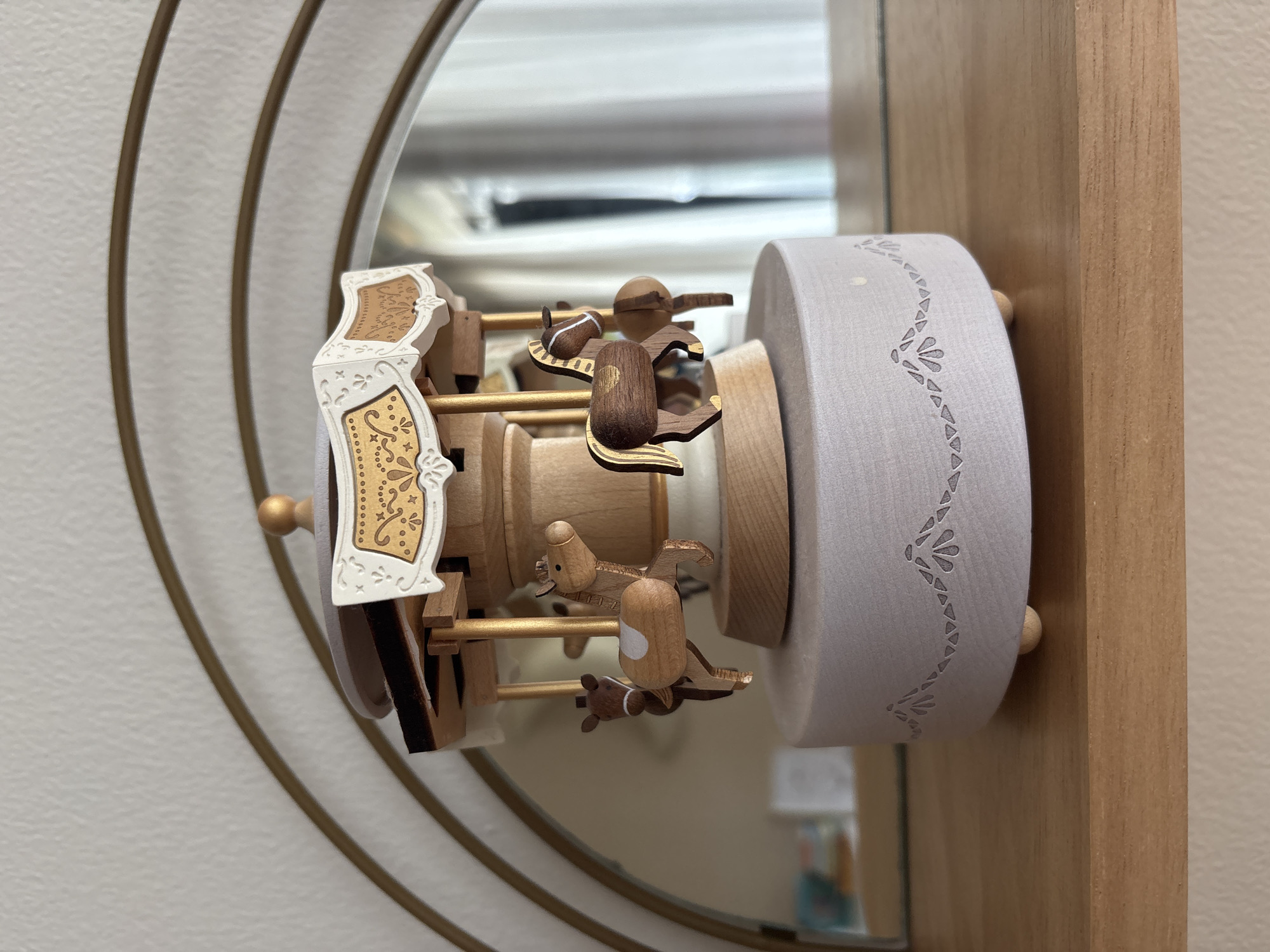 A wooden carousel music box, featuring three horses with gold accents, sits on a light purple base decorated with a patterned border. The music box is placed on a wooden shelf, against a wall with a large, arched mirror behind it. The mirror reflects a window with white curtains. The scene is indoors, and there is no indication of the city of Dubai or the United Arab Emirates.