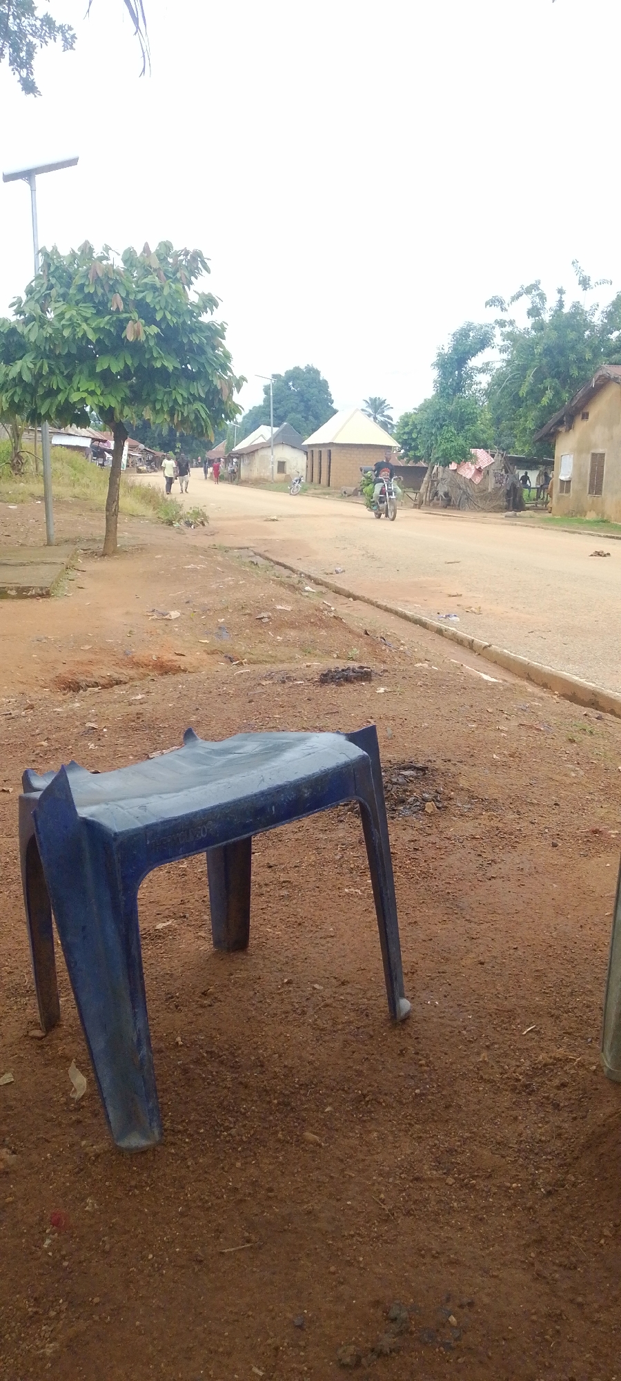 This media file captures a scene in Baissa, Nigeria, depicting a rural roadside setting. In the foreground, a blue plastic stool is placed on reddish-brown dirt. The stool is a simple, four-legged design, appearing somewhat weathered.
The background reveals a dirt road with a few people walking and a motorcycle carrying goods. The road is bordered by simple buildings, some appearing to be made of mud or earth with thatched roofs, while others have more conventional construction with light-colored walls and darker roofs. A few trees are scattered throughout the scene, with one prominently featuring green foliage in the mid-ground. A solar-powered street lamp is visible on the left side of the road.
The time of day appears to be overcast or late afternoon, as the sky is bright white, suggesting diffused light. The weather seems calm. The overall impression is one of a typical day in a rural Nigerian community, with everyday activities taking place along the road. There is no discernible text visible in the image.