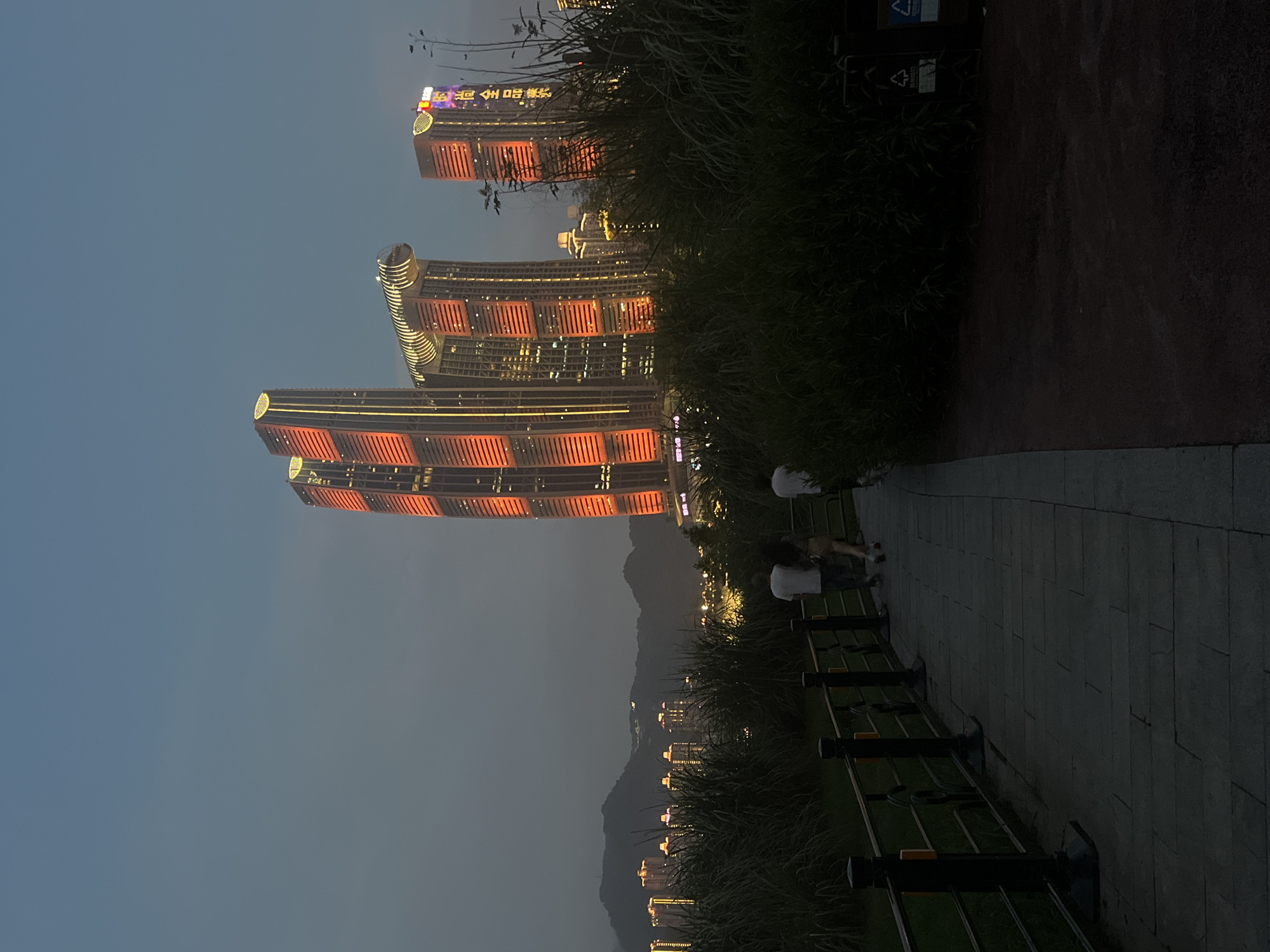 In the city of Jiangbeicheng Street, China, at dusk, a paved walkway winds through dense vegetation. Three individuals, two men and one woman, stroll along the path, their backs to the viewer. The male on the left wears a white t-shirt, the woman a light-colored top and skirt, and the male on the right another white t-shirt. In the background, modern skyscrapers illuminate the twilight sky with vibrant orange and yellow lights. Distant mountains are silhouetted against the fading light. A dark metal railing runs alongside the walkway, with black posts supporting it. To the right of the path, a red-surfaced area and dark green foliage dominate the foreground.