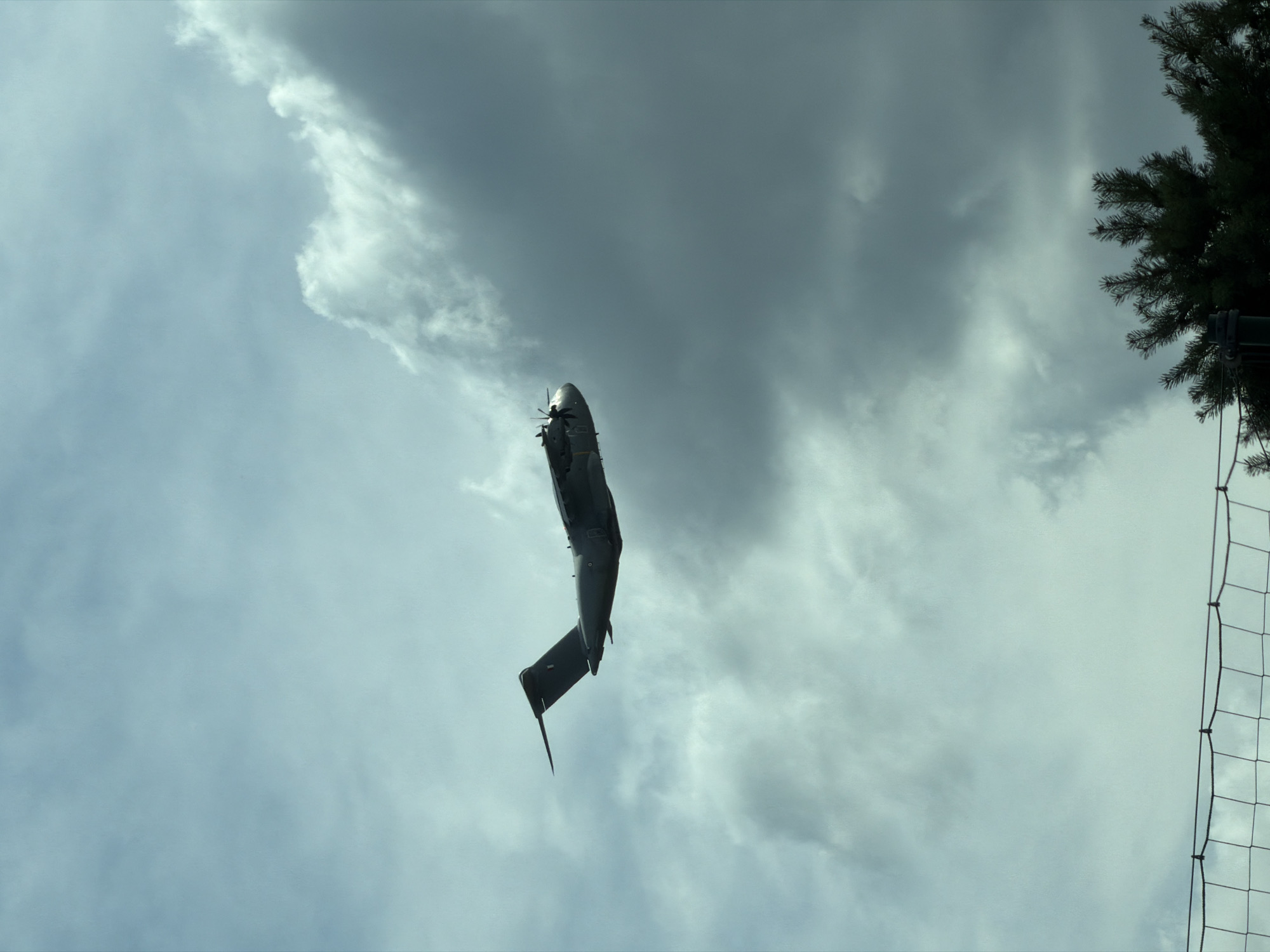 A large, dark gray military transport aircraft, possibly an Airbus A400M, is captured in mid-flight against a cloudy sky. The aircraft is angled slightly upwards, with its four propellers visible in the foreground on the left side. The tail of the aircraft is on the right side of the image, featuring a dark blue or black insignia. The sky is filled with dramatic, layered clouds, ranging from light gray to dark, stormy hues, suggesting an overcast or approaching weather system. In the bottom right corner of the image, the dark green needles of a coniferous tree are visible, partially obscuring the lower part of the frame. A section of a dark net fence, possibly a sports field boundary, is also visible along the bottom edge of the image. The scene appears to be a snapshot of the aircraft in flight over a general area that could be Vélizy-Villacoublay, France, given the context provided.