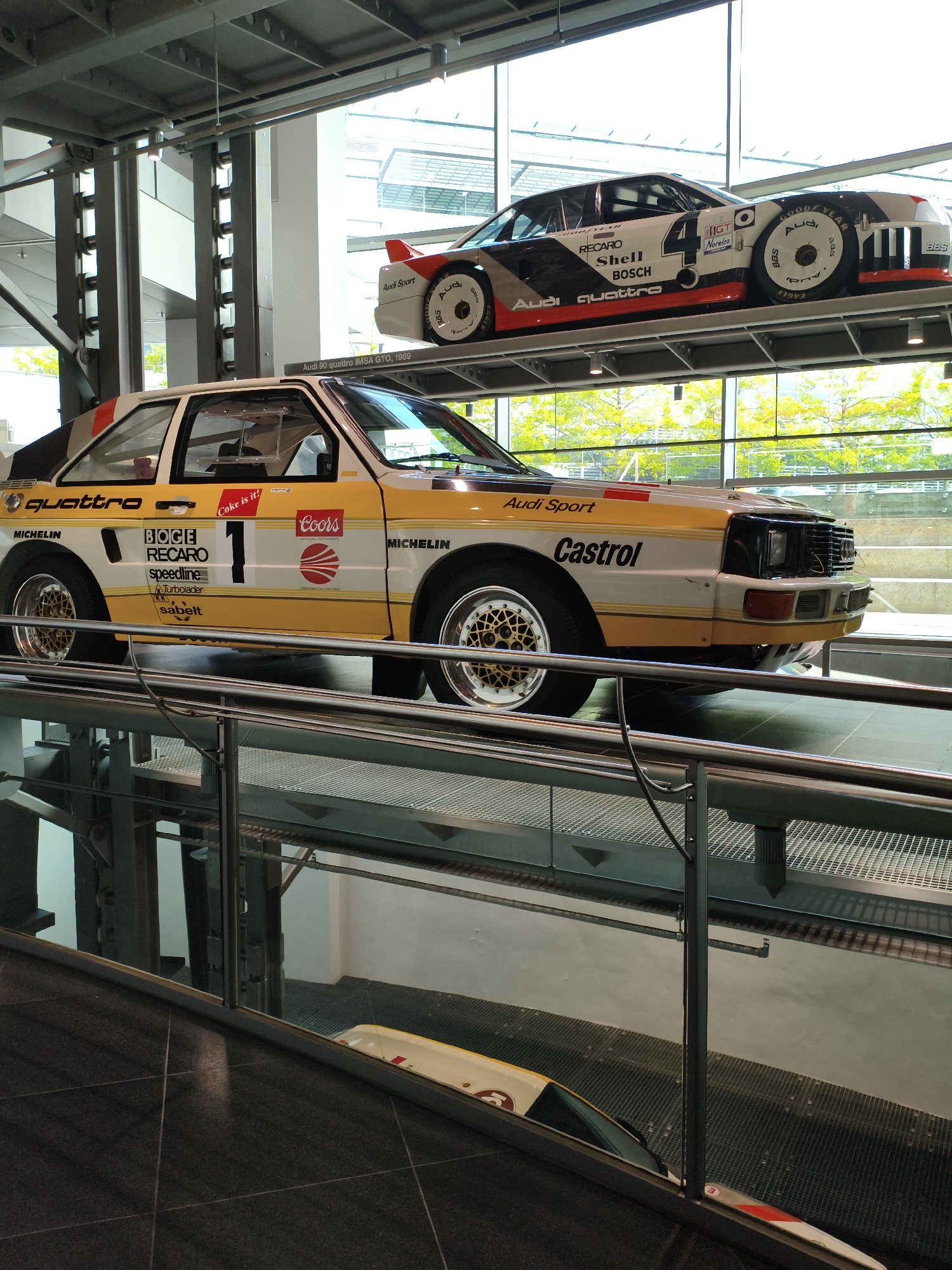 The image shows two Audi rally cars displayed on elevated platforms in a modern, well-lit building with large glass windows. The car in the foreground is a white and yellow Audi Sport quattro with various sponsor logos, including "quattro," "Michelin," "BOGE," "RECARO," "SPEEDLINE," "TurboIsolator," "Sabelt," "Coke is it!", and "Coors." It has the number "1" prominently displayed on its side. The car in the background, partially visible, is a black, white, and red Audi 90 quattro IMSA GTO, 1989, also featuring sponsor decals such as "RECARO," "Shell," "Bosch," "Audi quattro," and "Goodyear." The cars are positioned on metal display structures. The background reveals a cityscape with trees and modern architecture visible through the windows. The overall impression is that of an automotive museum or exhibition space, likely in Ingolstadt, Germany, the headquarters of Audi.