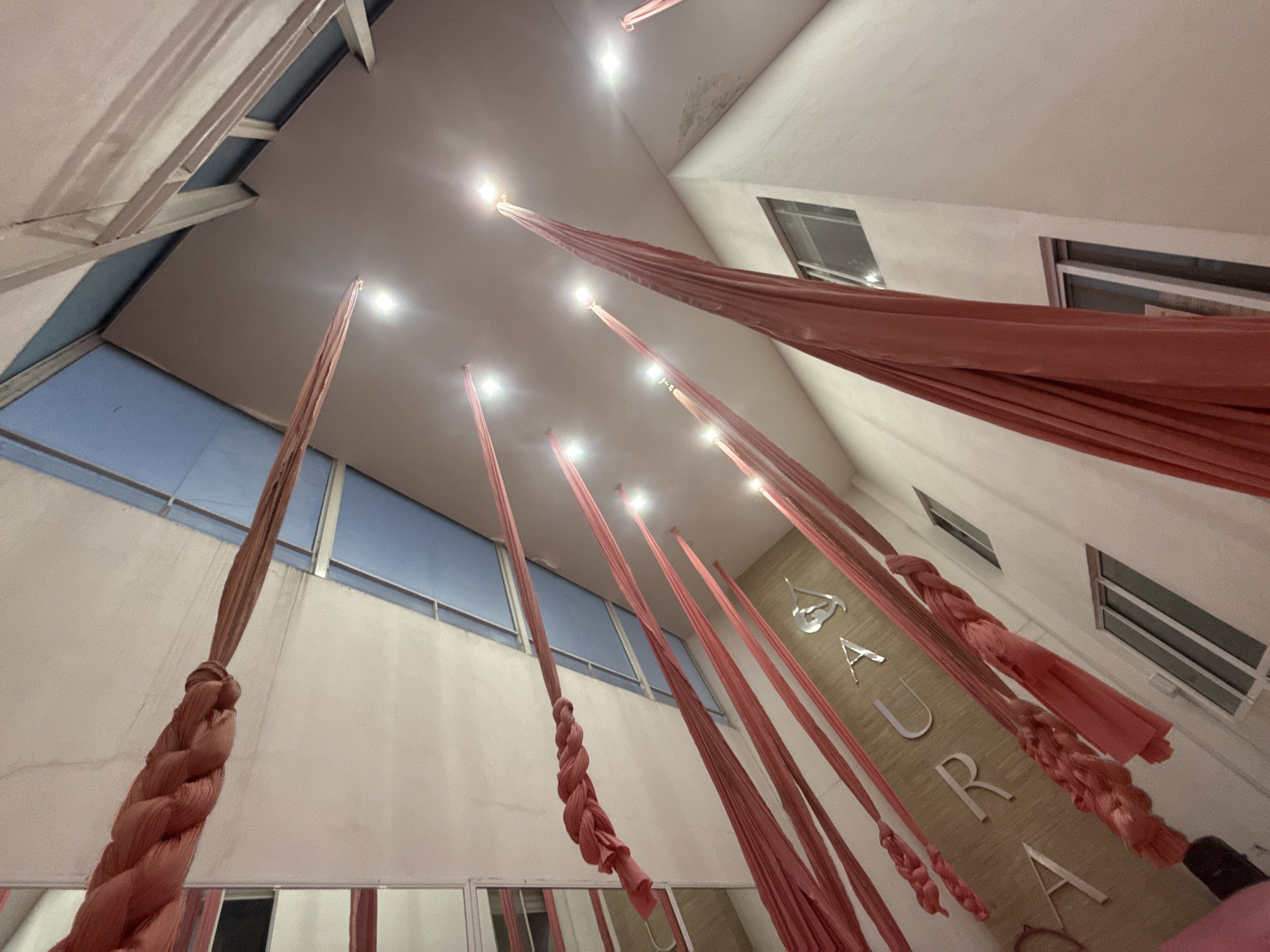The image shows an interior space in San Andrés Cholula, Mexico, where aerial silks are suspended from the ceiling. The building has white walls and ceiling, with windows visible on multiple walls. Several vertical pinkish-red silks hang from the ceiling, illuminated by bright lights at the attachment points. The silks are gathered and knotted at varying heights. Silver lettering that spells out "SAURA" is affixed to one wall. Reflections of the silks and the environment are visible in what appears to be mirrors along the bottom edge of the frame. The overall perspective is angled upwards.