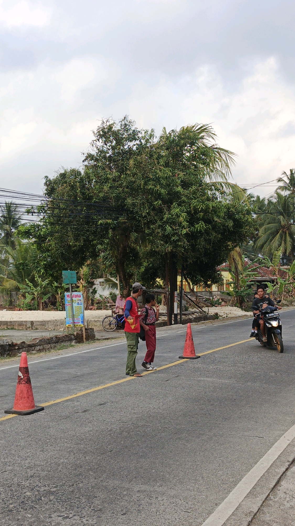 In Cibenda, Indonesia, a man in a red shirt and green pants stands on the asphalt road, facing a younger person in red pants and a patterned shirt. The man appears to be speaking to the younger person, holding a yellow object. Two orange traffic cones are visible on the road, one in the foreground and another further back. A motorcycle with two occupants passes by on the right side of the road. In the background, trees and houses are visible, with a cloudy sky overhead. A sign with text and images is also present on the left side of the road.
