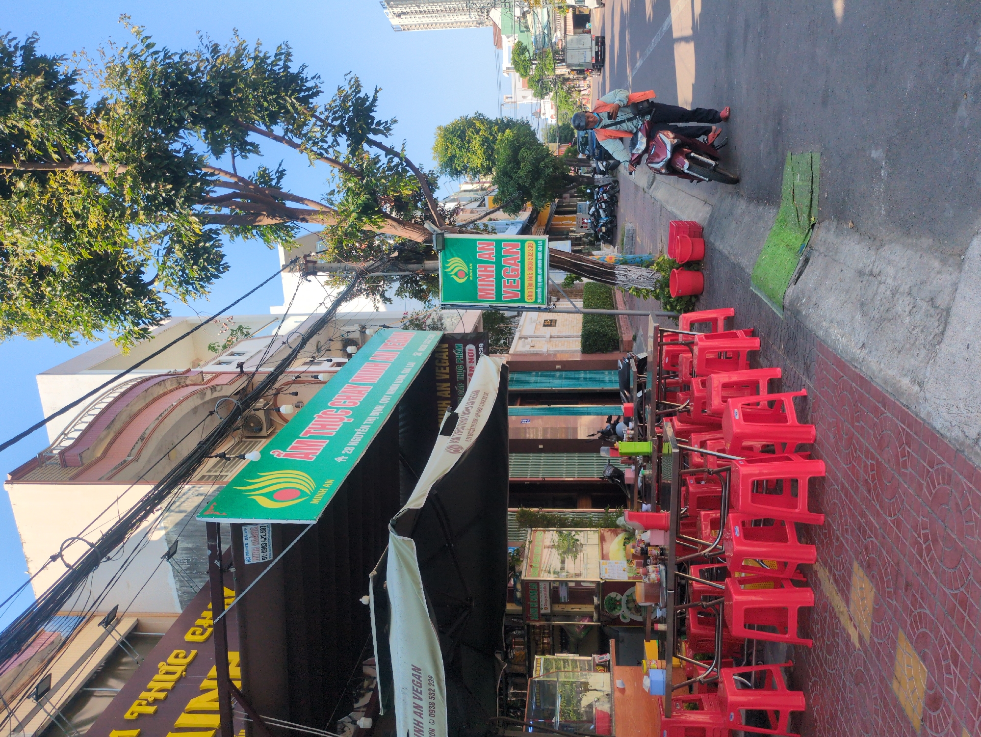 The image depicts a street scene in South Quy Nhon Ward, Vietnam, featuring a vegan restaurant.
The restaurant, "Minh An Vegan," has a green sign with its name and contact information. Several red plastic chairs are stacked on the sidewalk in front of the restaurant. There is an awning over the outdoor dining area.
In the background, a man on a motorbike is on the street.