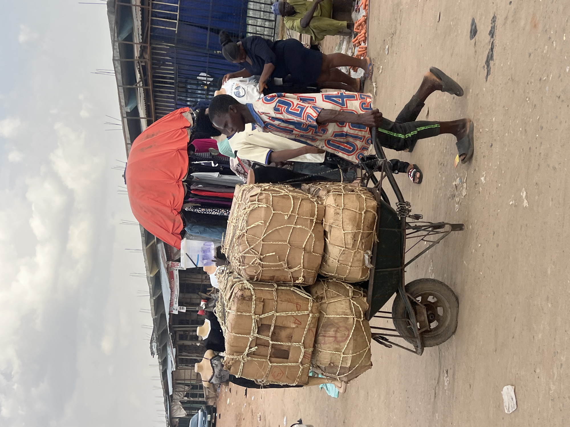 In Oko Amakom, Nigeria, a man wearing a patterned shirt and dark pants pushes a heavily laden two-wheeled cart along a concrete surface. The cart is stacked with six large, rectangular packages, each wrapped in brown material and secured with netting. An open red umbrella is positioned near the cart, likely to provide shade for items displayed for sale nearby, including clothing hanging on racks. Other people are in the background, including a woman in a dark dress and another individual in light-colored clothing. Buildings with corrugated metal roofing form part of the backdrop to the scene. The sky is overcast.