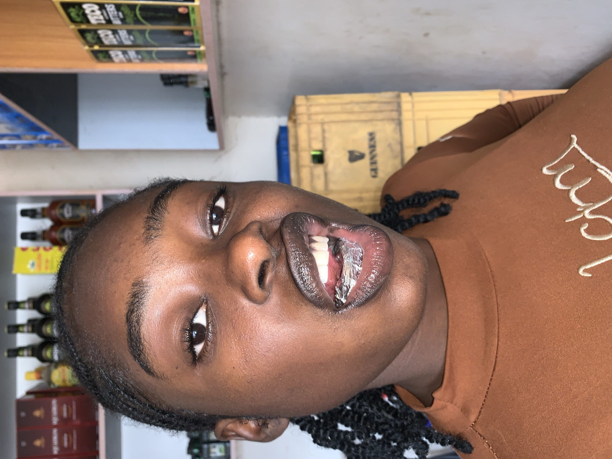 A close-up, eye-level portrait depicts a person with dark skin and braided hair looking directly at the camera. The individual's mouth is open, revealing white teeth and a silver foil-wrapped object positioned between the lower lip and lower teeth. The person wears a brown, high-necked, long-sleeved top with partial white embroidery on the chest. The background, indicative of an indoor retail or storage setting, features multiple shelves. The shelf to the left displays red-boxed items, a yellow-labeled bottle, and several dark bottles with green labels. The shelf to the right holds packages labeled "TECO BITTERS." Below this, two stacked wooden crates, the top one bearing a black "GUINNESS" logo, are visible against a light-colored wall. The scene is located in New Stadium, Nigeria.